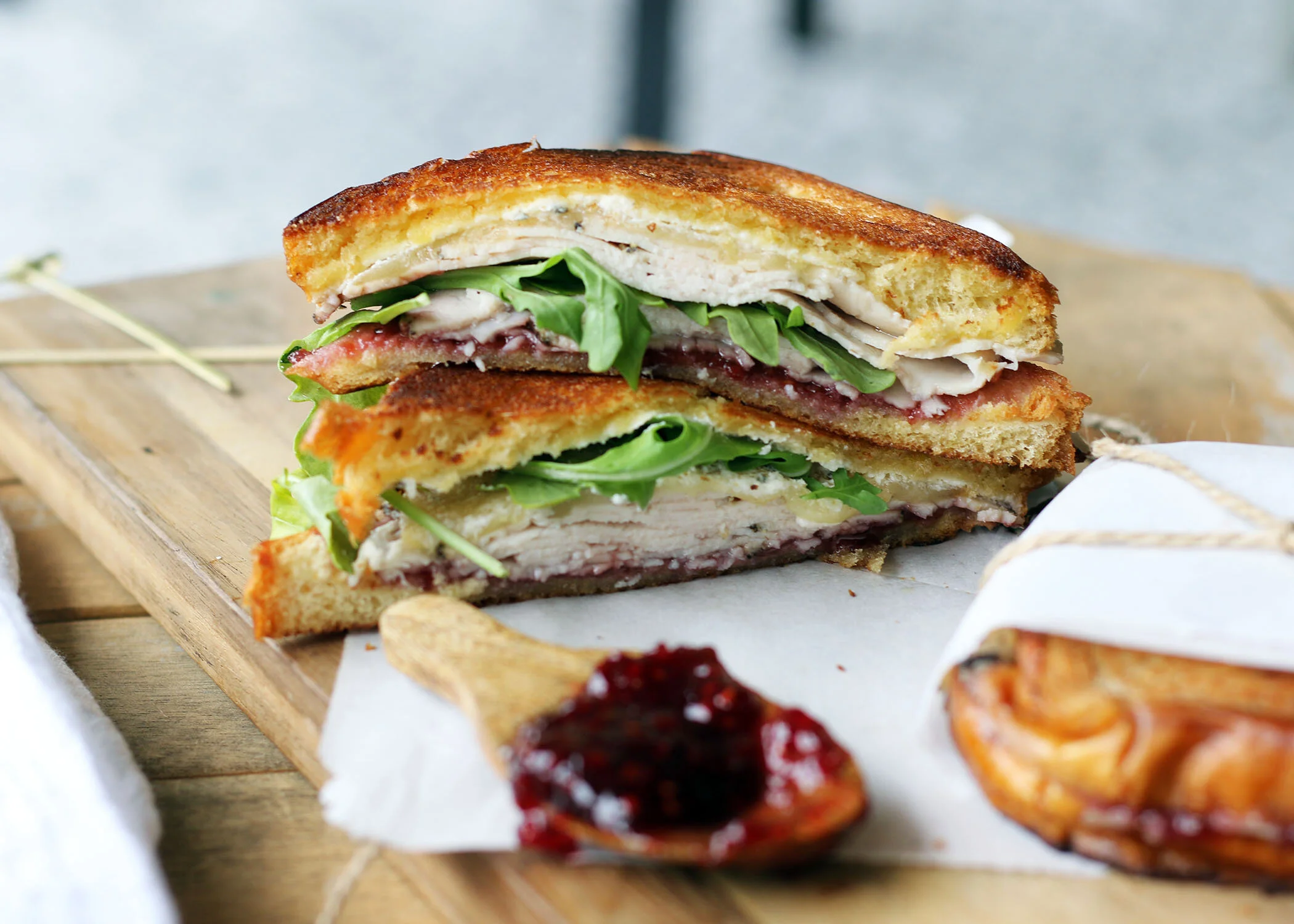 Gourmet sandwich with turkey, arugula, cheese, and cranberry sauce on toasted bread, served on a wooden board with a spoonful of jam.