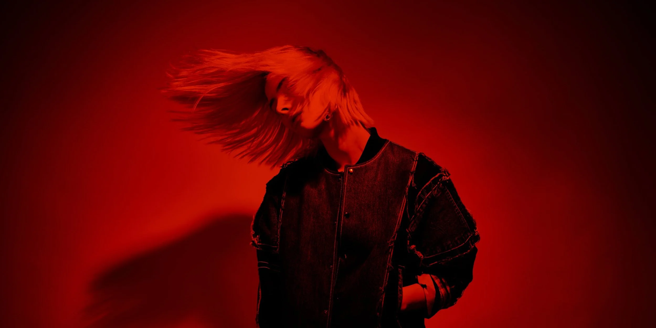 A young woman throws her head to her right side, with her long hair whipping around her face. She is cast in a strong red light.