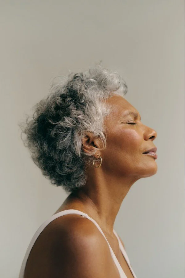 Side profile of an elderly woman with gray curly hair, closed eyes, and a peaceful expression, wearing a pink strap top.