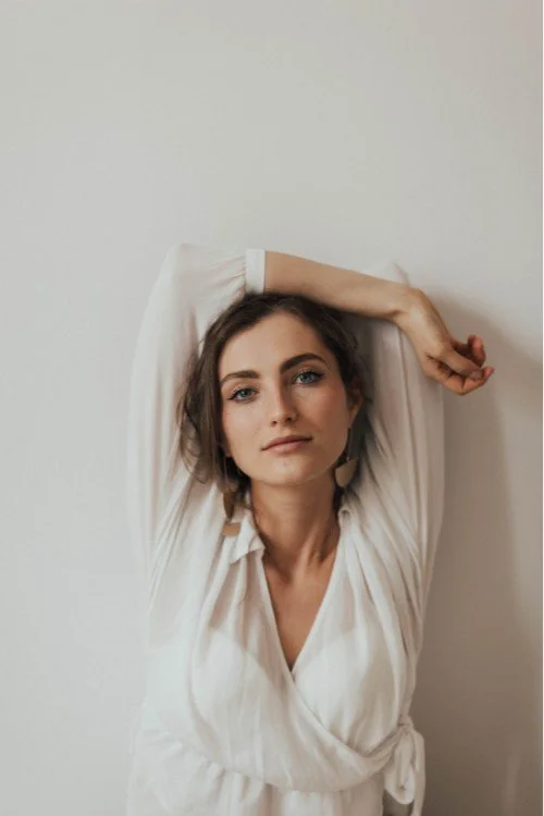 Young woman with brown hair and blue eyes wearing a white blouse, posing against a plain light-colored wall with her left arm behind her head.