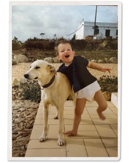 A young child laughing and hugging a light-colored dog outside on a tiled patio, with a background of rocks, plants, and a white building under a cloudy sky.