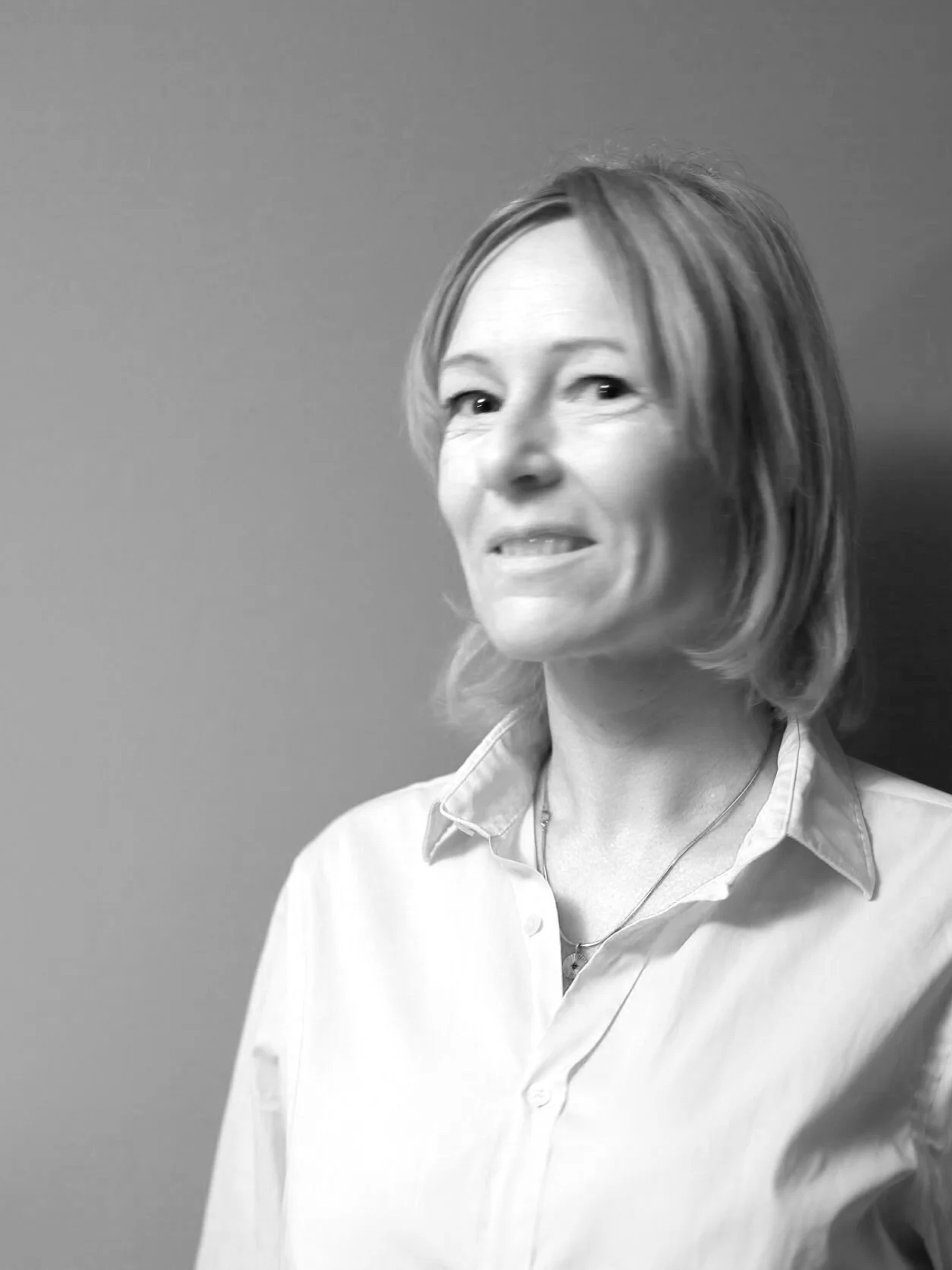 Zwarte en witte foto van een glimlachende vrouw met schouderlange haar, gedragen in een lichtkleurig blouse, zittend voor een effen achtergrond.