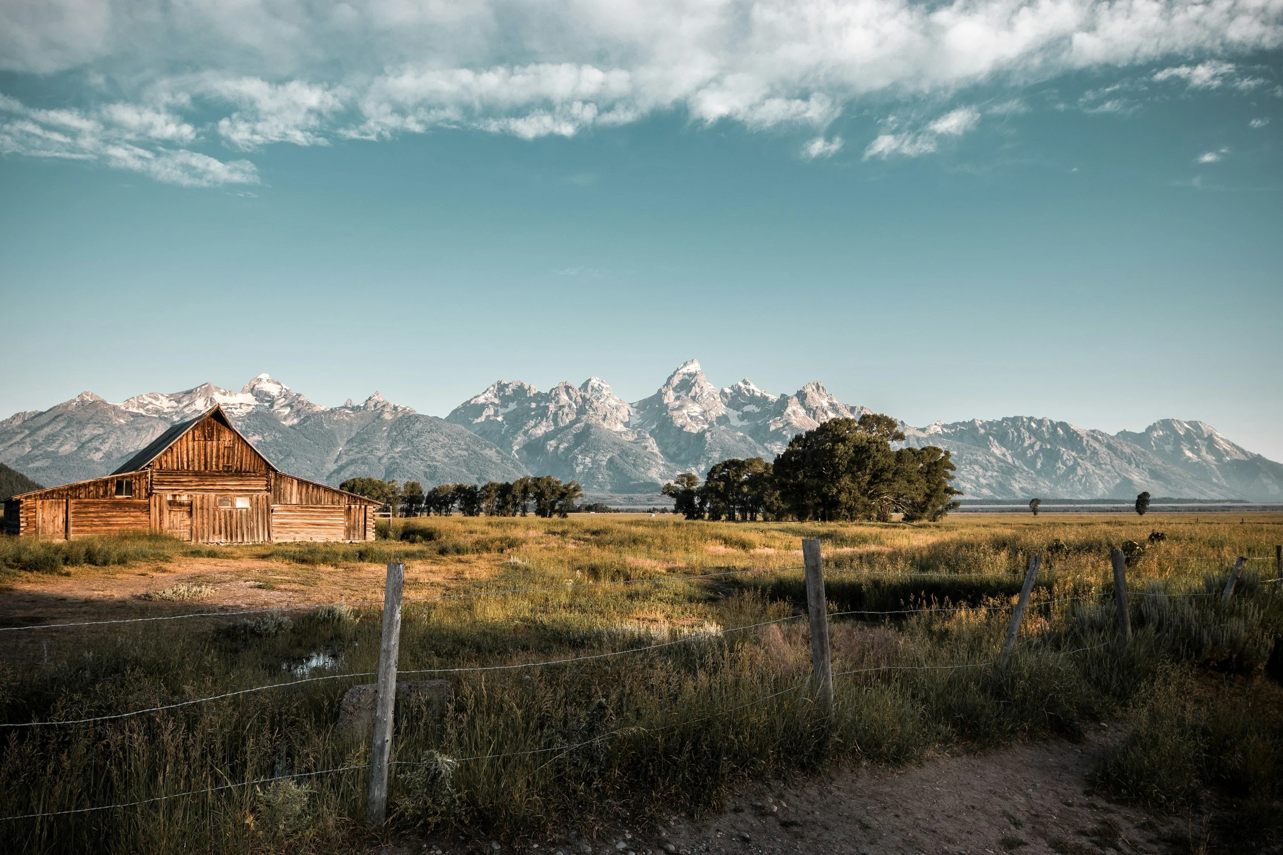 A rustic wooden barn in a grassy field with a mountain range in the background under a partly cloudy sky.