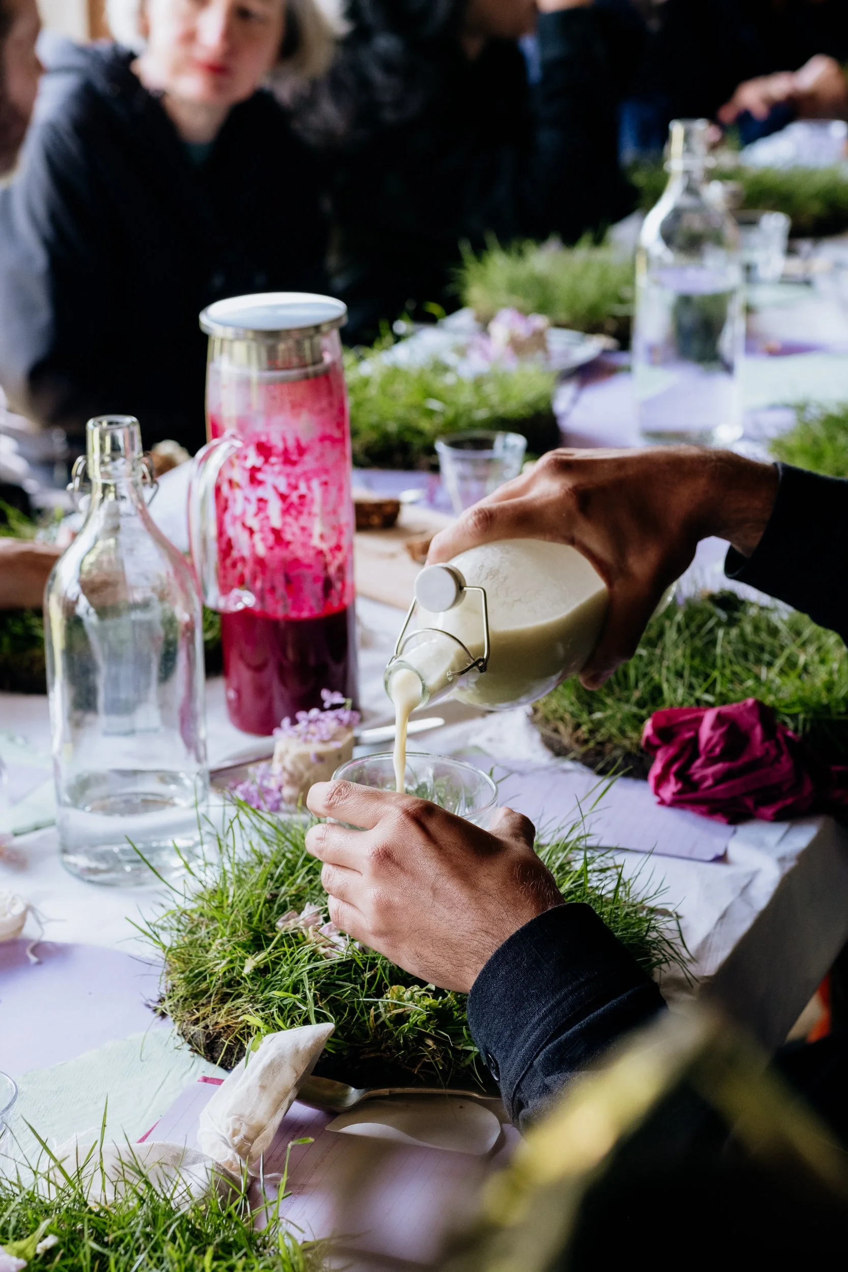 a close up of some milky liquid being poured from a glass bottle into a glass. There is a long table and set out for a meal and there is grass on the table. There is also a flask containing a beetroot coloured liquid.