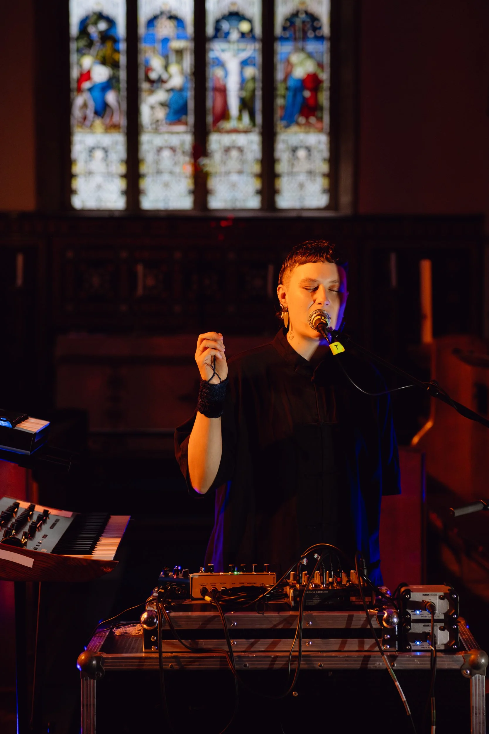 A person sings into a microphone in a church. There is a large stained glass window behind them.