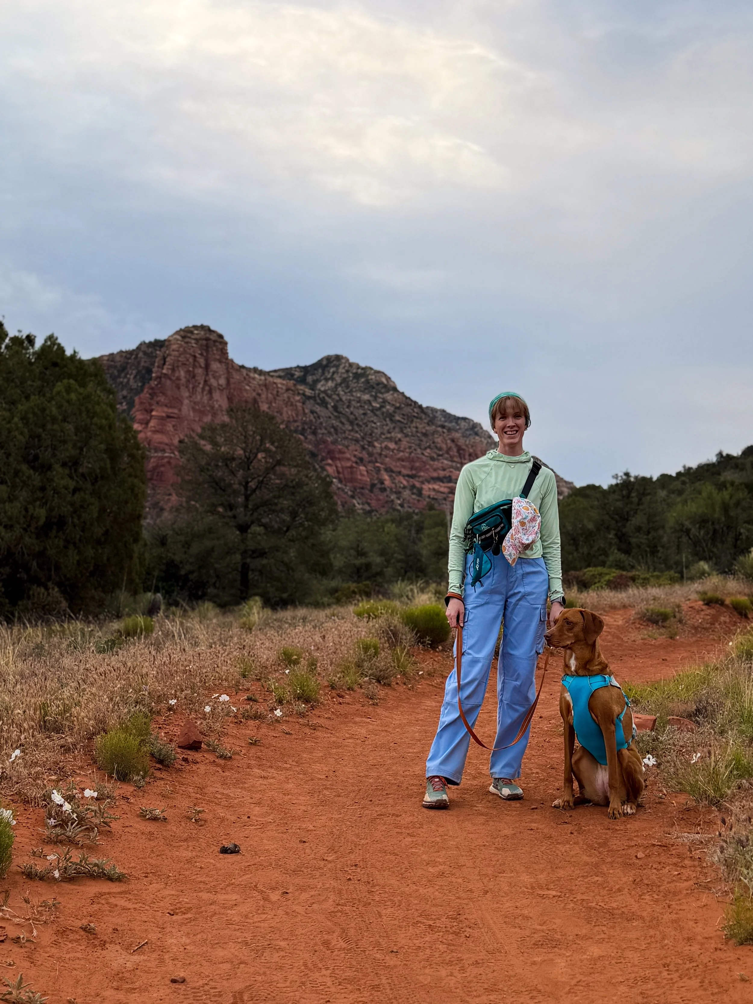 A woman in hiking gear standing on a dirt trail with her brown dog in a natural landscape with red rock formations and trees in the background.