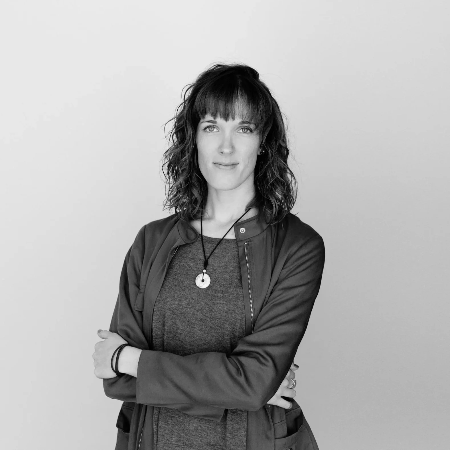 A woman with shoulder-length wavy hair, wearing a dark jacket and a necklace, standing with arms crossed against a plain light background.