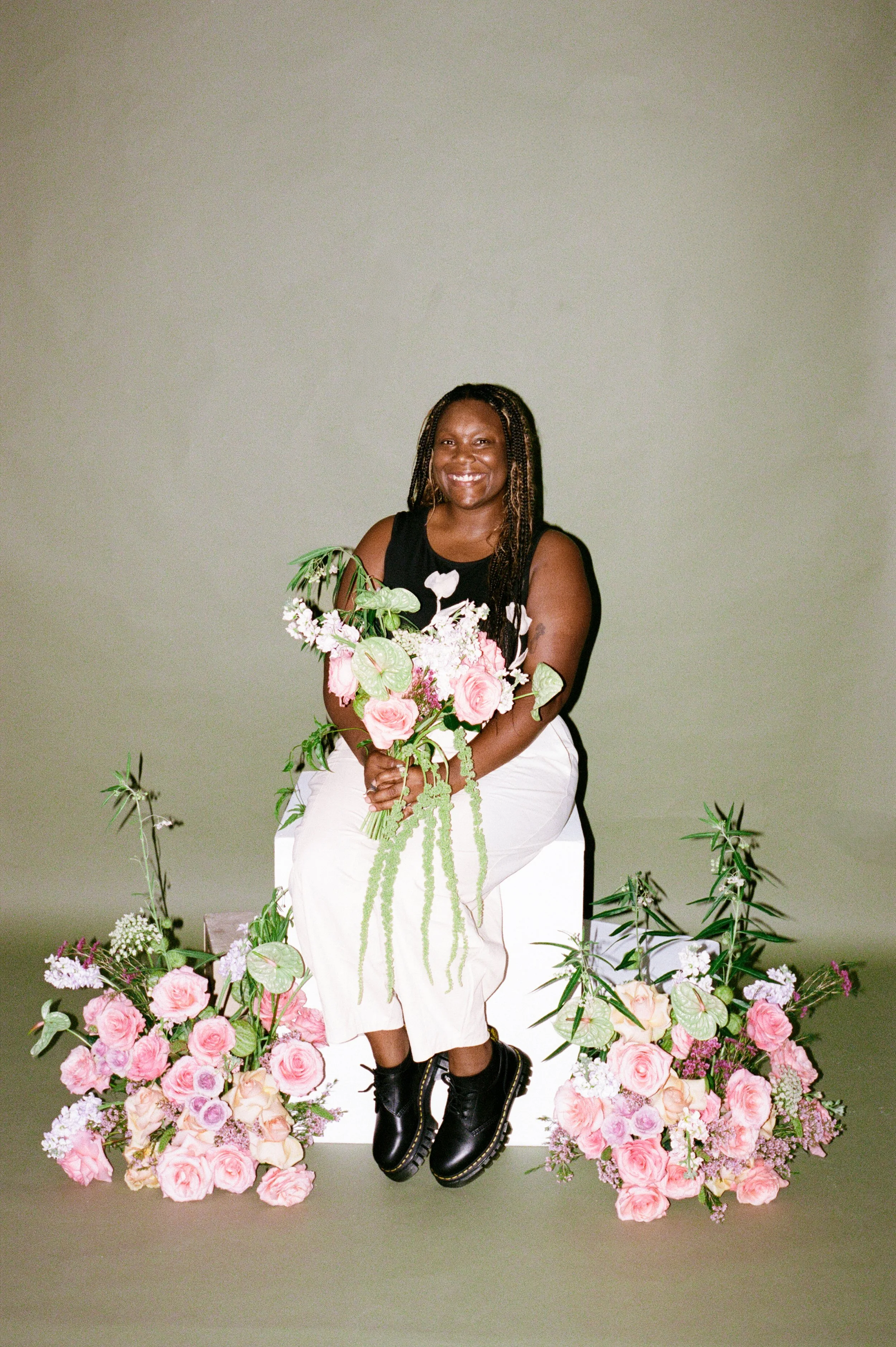 A smiling woman with dark skin and braids, sitting on a white block, holding a bouquet of pink and white flowers. She is dressed in a black sleeveless top and white pants, surrounded by pink floral arrangements and green foliage, with a plain light g