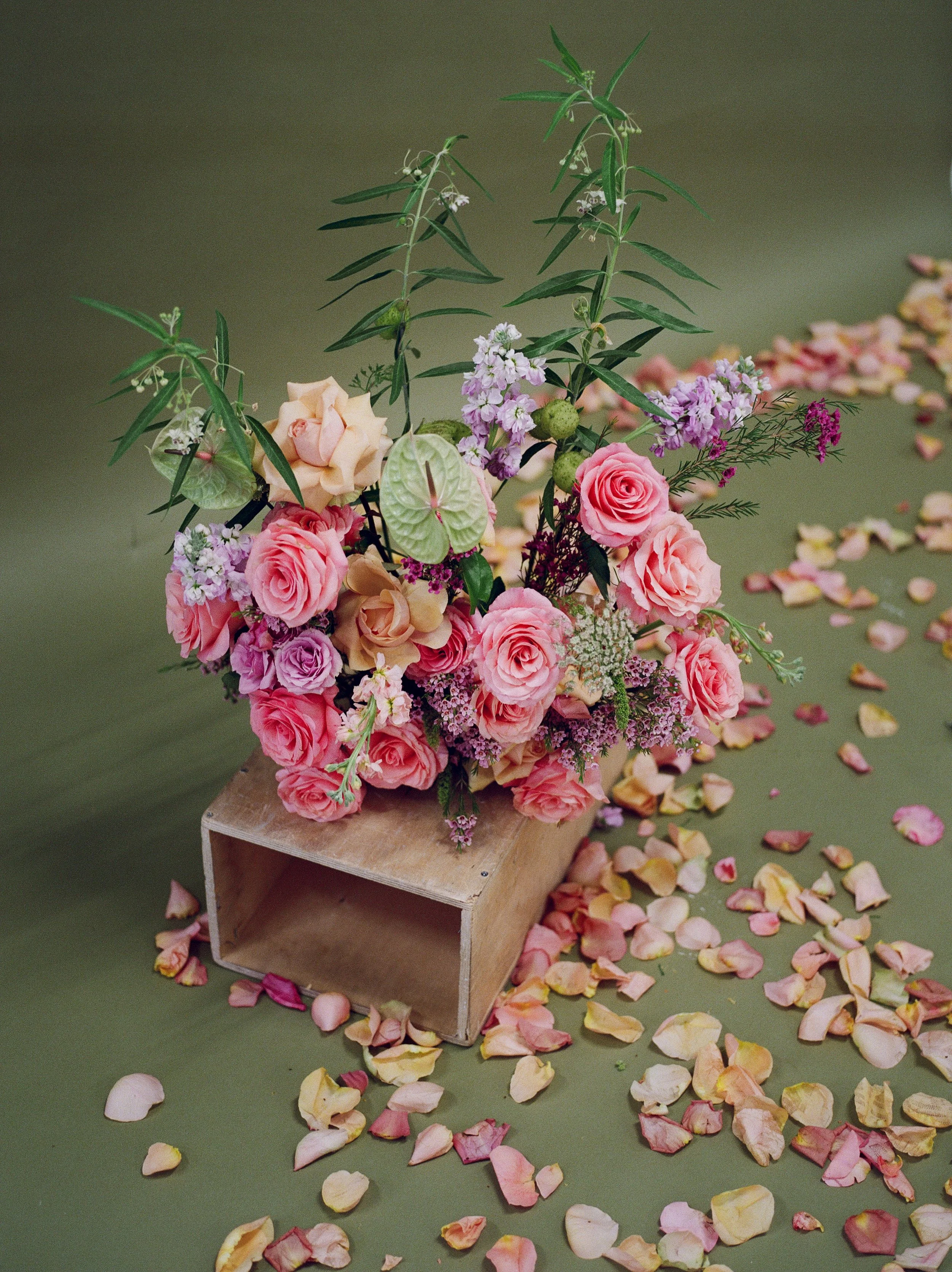 A floral arrangement of pink roses, purple and white smaller flowers, and green foliage in a vase, placed on small wooden block, with rose petals scattered around on green surface.