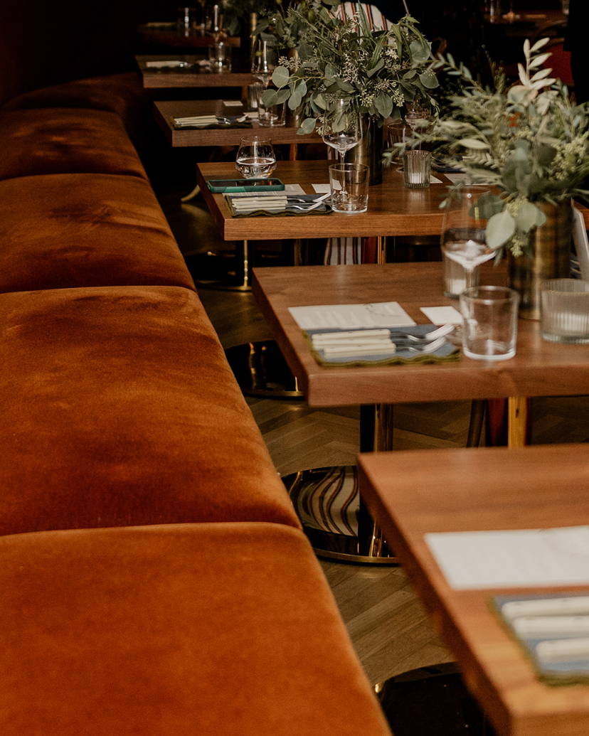 Empty restaurant tables with glass vases filled with green foliage, set with menus, glasses, and silverware, next to a long, orange velvet booth.