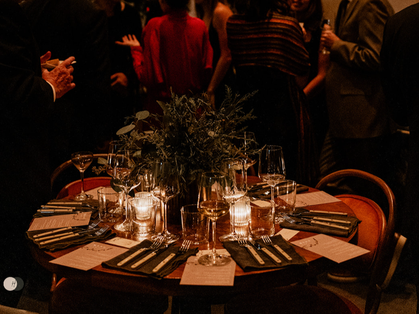 A round table set for a formal dinner with black napkins, silverware, glasses, and candles, topped with a floral centerpiece, in a dimly lit setting with people mingling in the background.