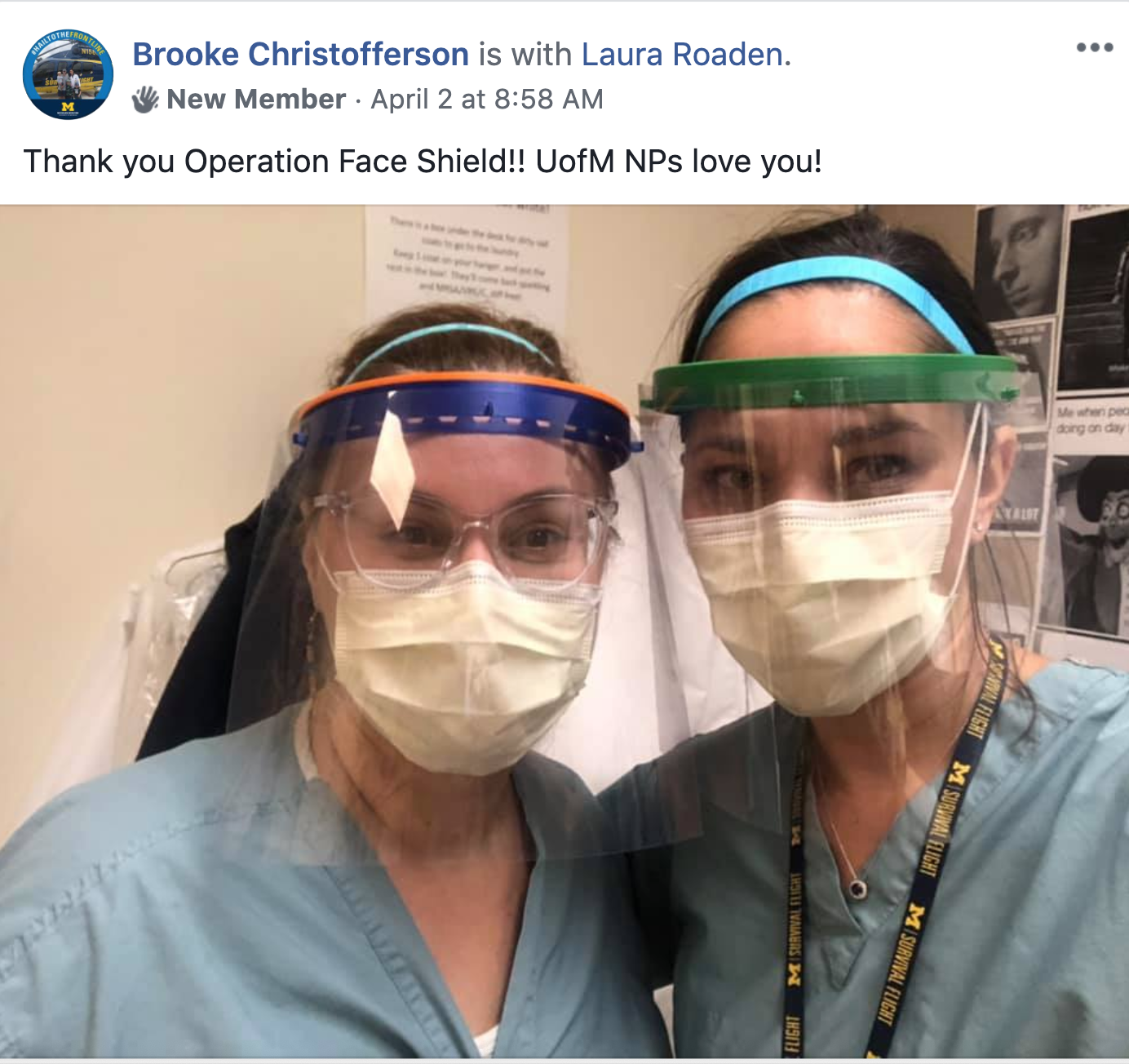 Two healthcare workers wearing masks, face shields, and scrubs pose for a selfie. Both are wearing protective gear and face masks, with posters visible in the background.