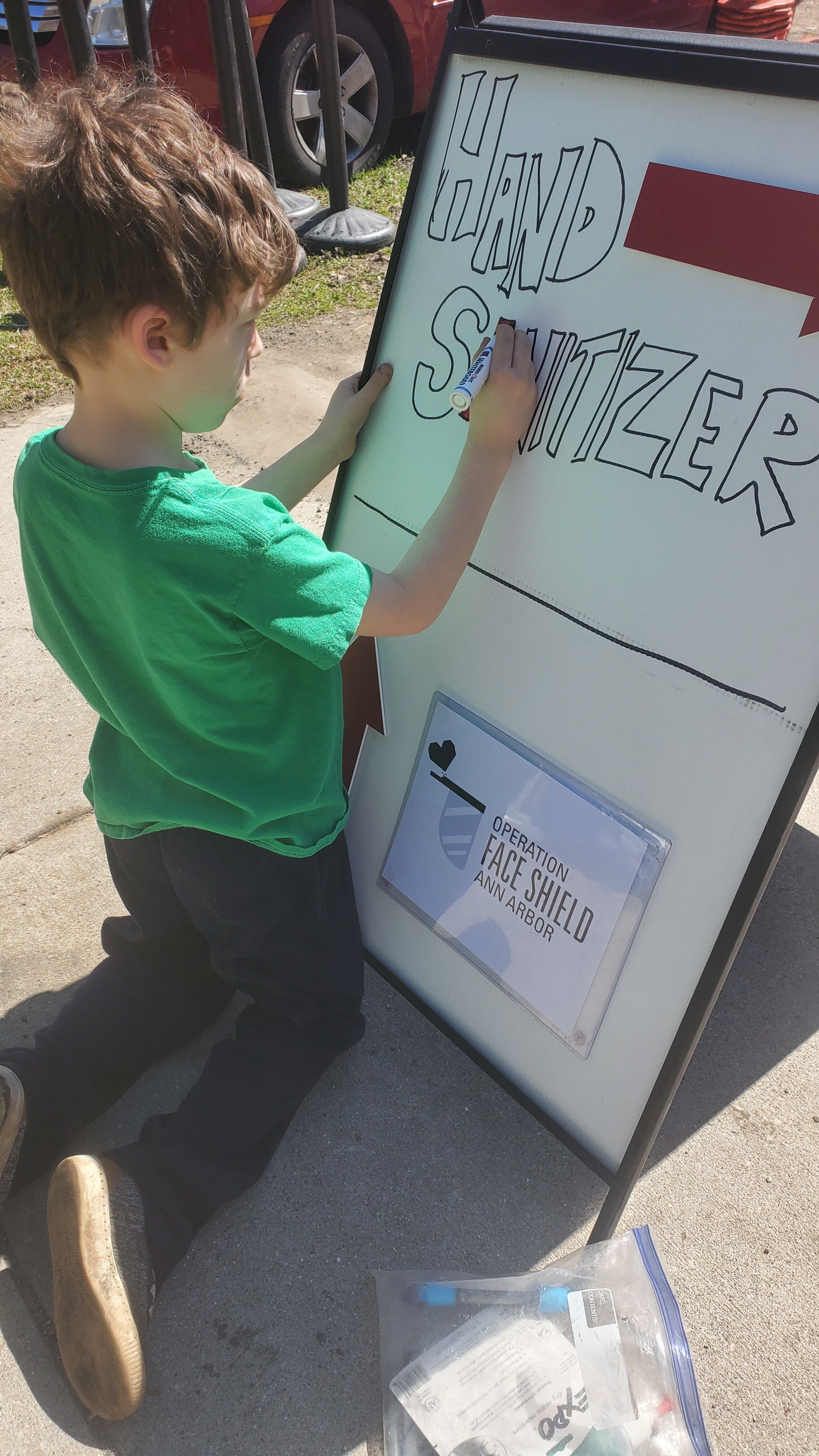 A young boy in a green shirt writing on a large whiteboard outside with a marker, titled "Hand Sanitizer" and a sign below reading "Operation Face Shield Ann Arbor."