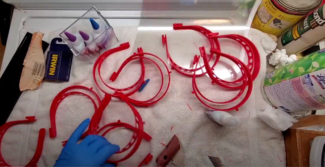A work surface with red embroidery hoops, a person's hand in a blue glove, and various art supplies including a container of glue, scissors, and rolls of tape.