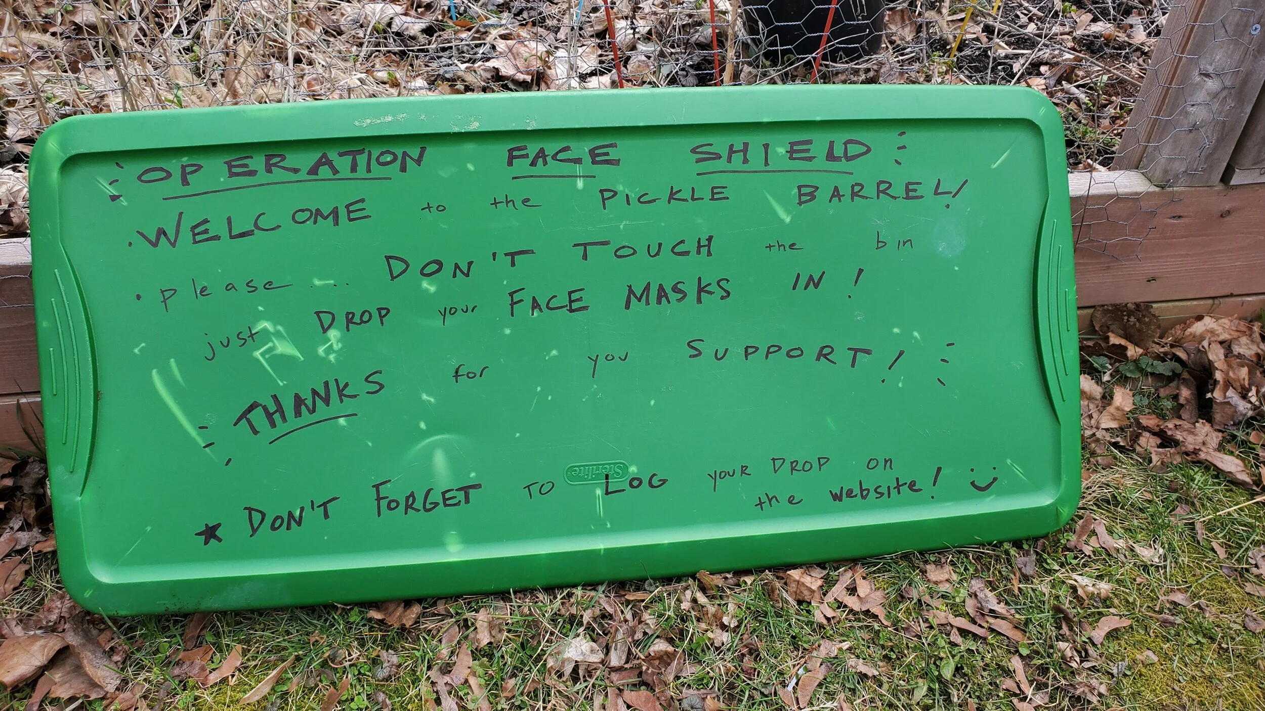 Green plastic sign with handwritten instructions for operation face shield, asking visitors to support by not touching the bin, dropping face masks in, and logging their drop on the website, placed on grass and leaf-covered ground next to a wooden fe