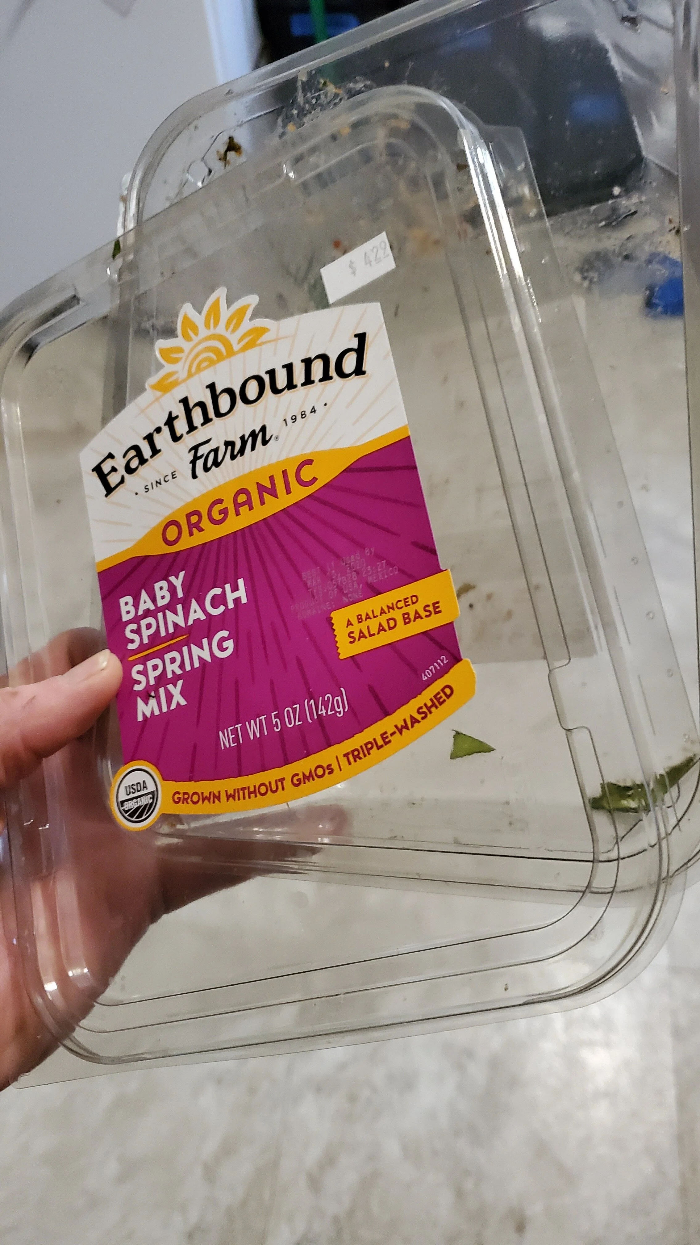 A person holding a plastic container with a label that reads "Earthbound Farm Organic Baby Spinach Spring Mix." The container is empty except for a few leaves inside, and is being held over a gray background surface.