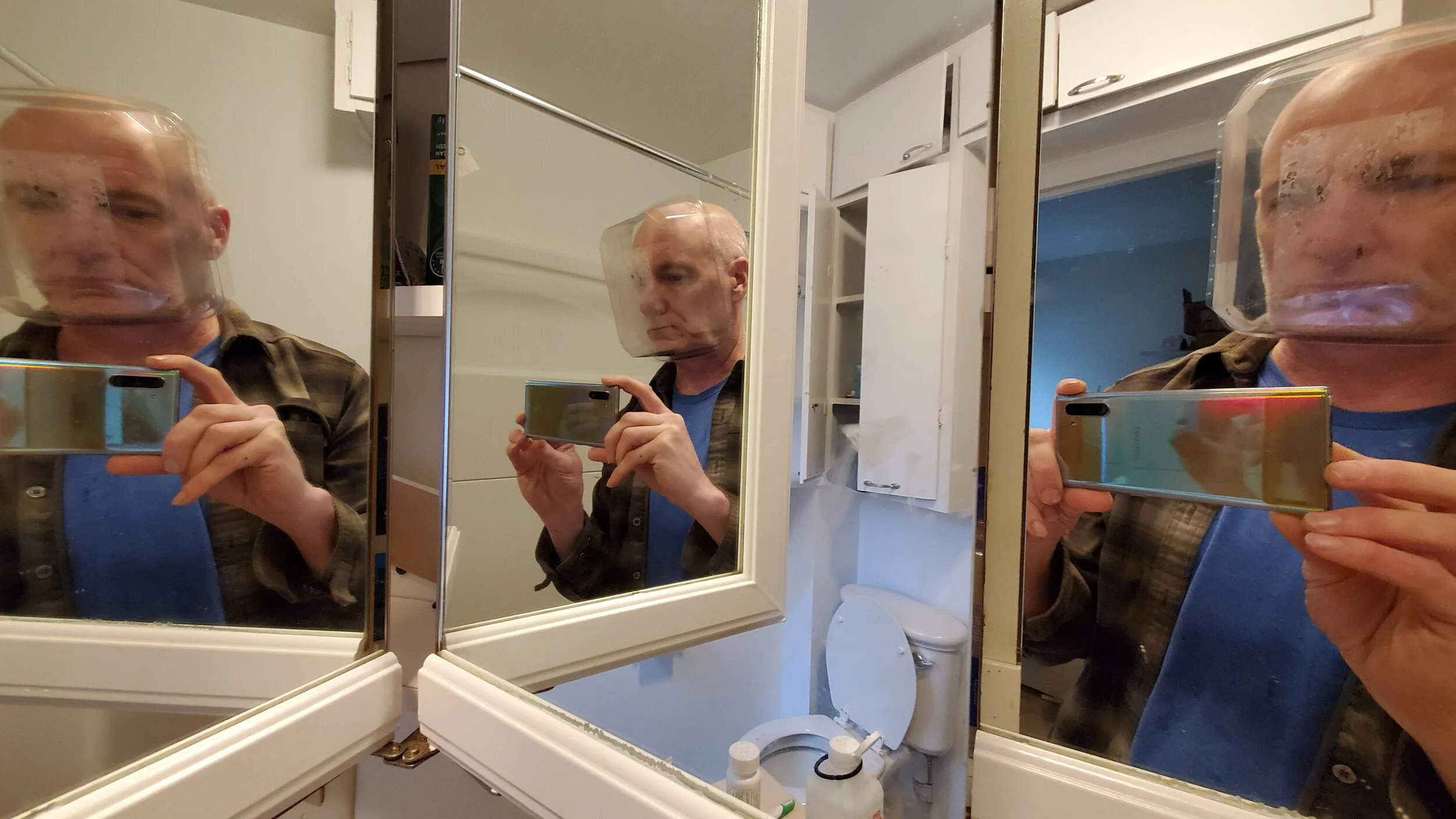 A man is taking a mirror selfie in a bathroom, wearing a clear plastic face shield, a blue shirt, and a brown plaid shirt. The mirror reflects multiple angles of the scene, including the toilet and bathroom cabinets in the background.