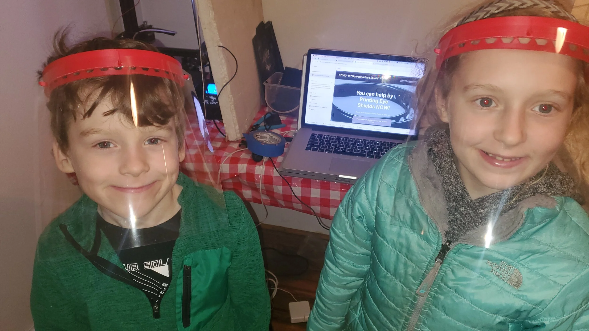 Two children, a boy and a girl, are wearing red face shield frames on their heads, smiling at the camera. Behind them, there is a laptop on a red and white checkered table with a webpage about face shields open.