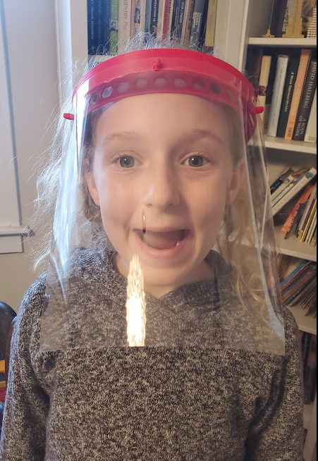 A young girl wearing a face shield with curly blonde hair, smiling in front of a bookshelf.