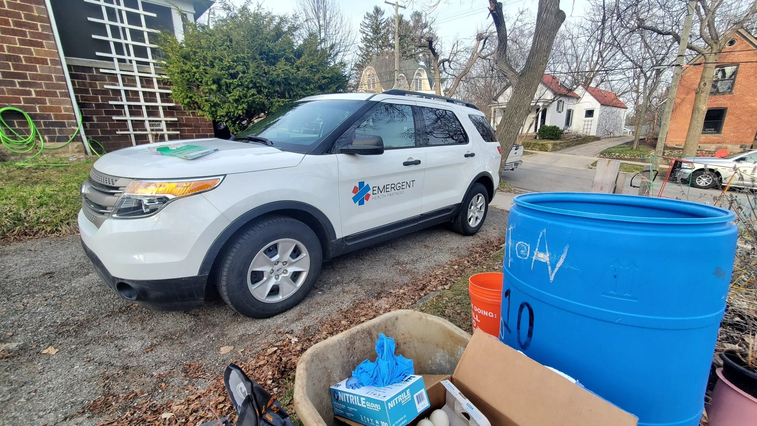 A white car with the logo 'Emergent Health Partners' parked on a gravel driveway, with various supplies including a large blue barrel, a box of nitrile gloves, and other items on a wheelbarrow in the foreground, and houses and trees in the background