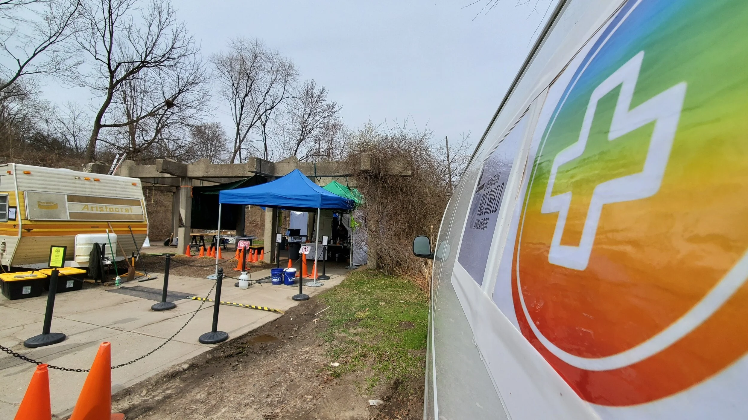 Outdoor COVID-19 testing site with tents, cones, and a mobile medical van, with trees and a concrete structure in the background.