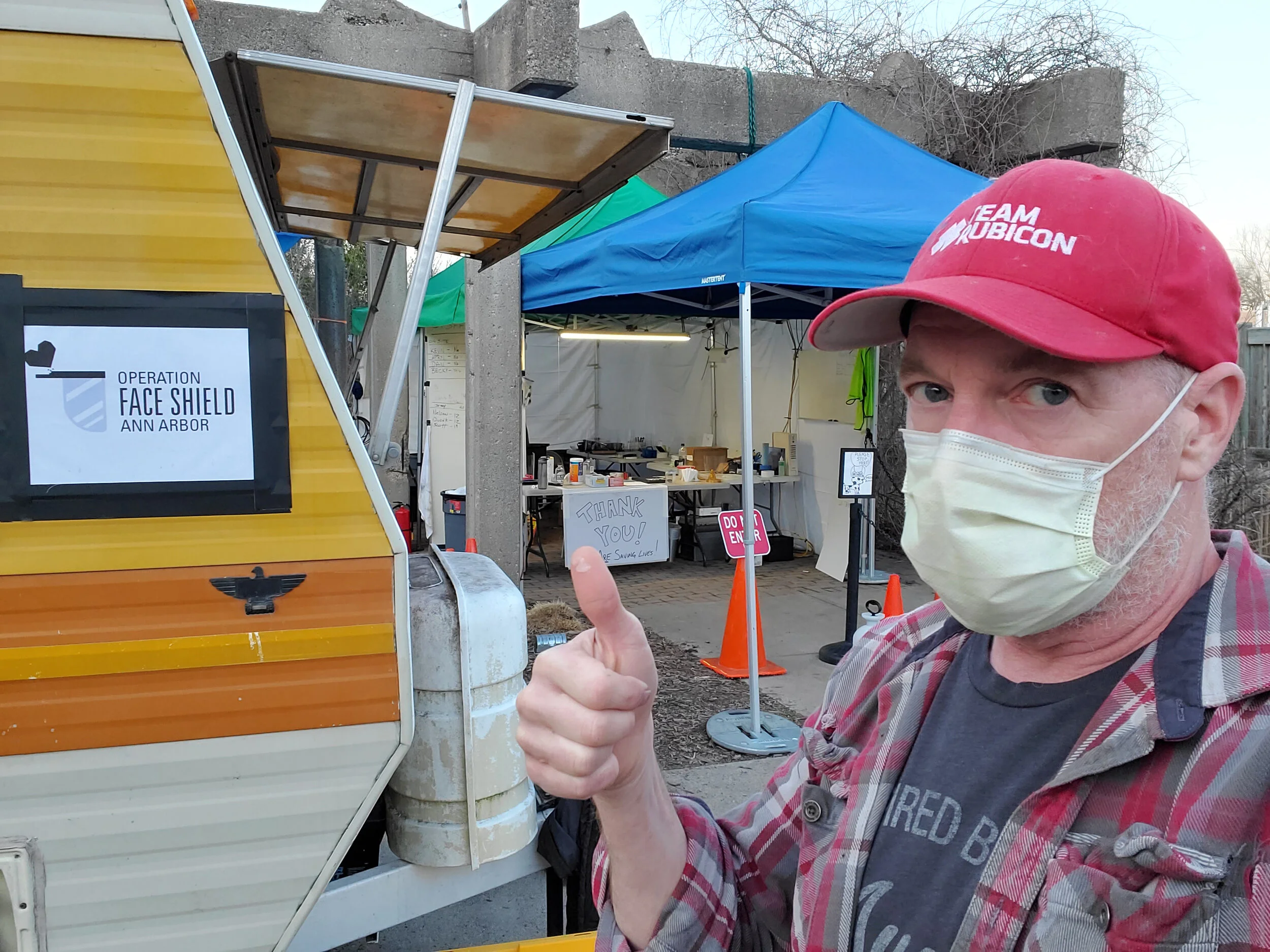 A man wearing a red cap and a face mask giving a thumbs-up in front of a food truck with a sign for Operation Face Shield Ann Arbor and an outdoor event area with blue and green tents.