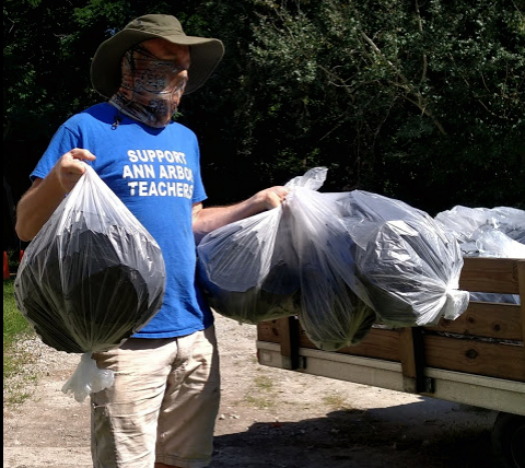 Person wearing a blue T-shirt that says support Ann Arbor teachers, a wide-brimmed hat, and a face covering, holding two trash bags filled with leaves or yard waste, standing outdoors next to a wooden cart filled with more trash bags.