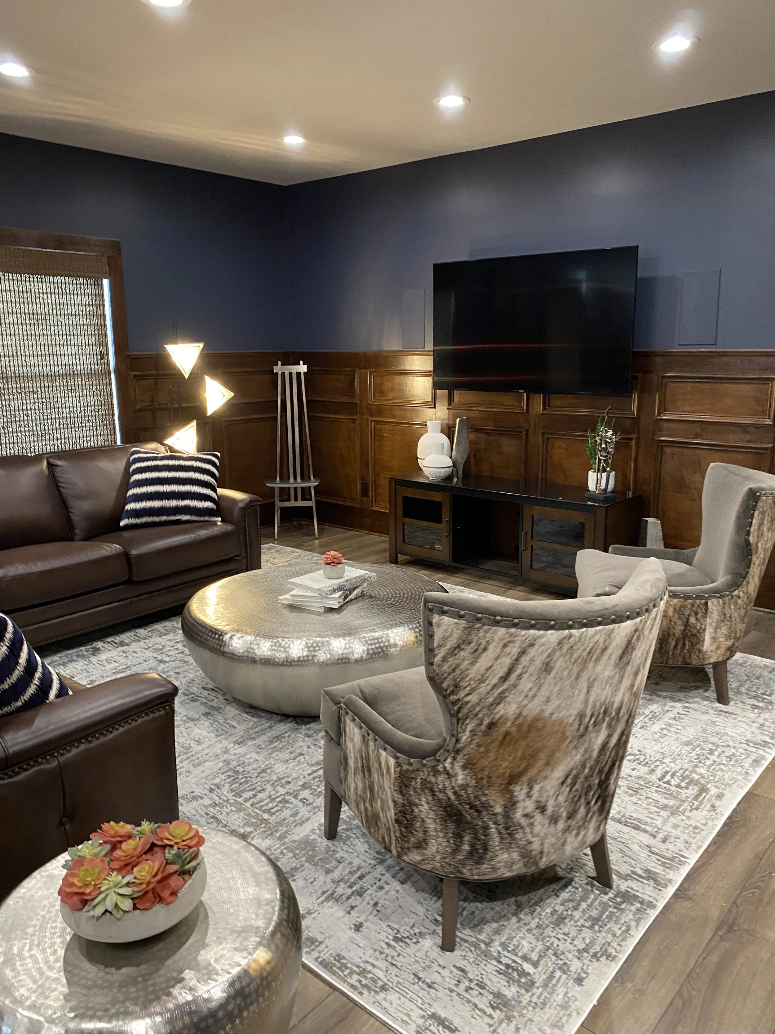Living room with a dark gray wall, wooden paneling, and ceiling lights. Includes a brown leather sofa, patterned armchair, coffee table with a floral arrangement, TV on a black stand with decorative vases and plants, and a tall floor lamp.