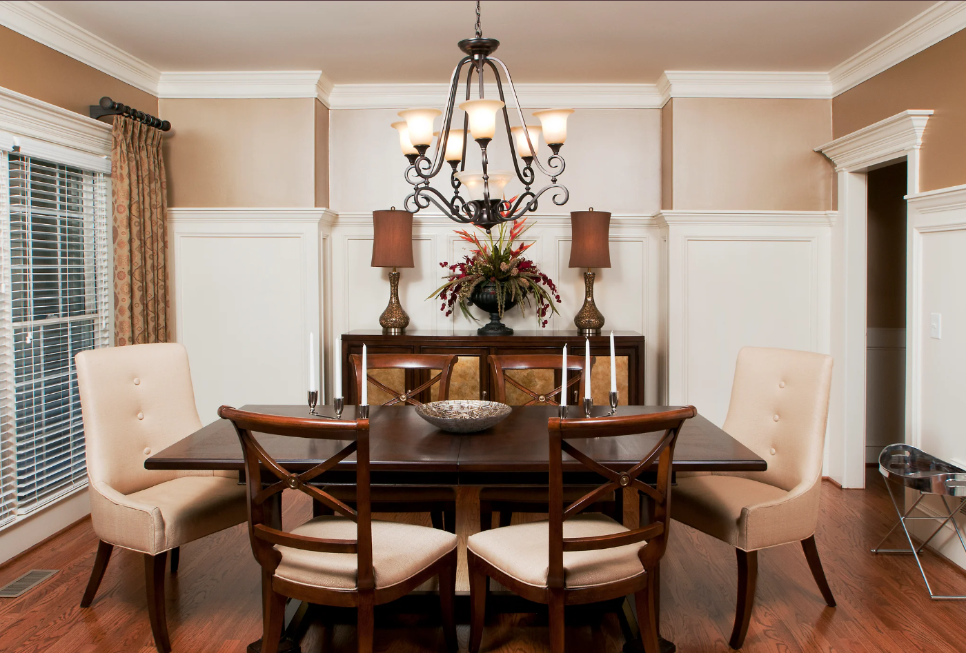 A dining room with a dark wood table, six chairs with beige cushions, a chandelier hanging above, and a sideboard with decorative lamps and a floral arrangement. There are windows with blinds and curtains on the left.