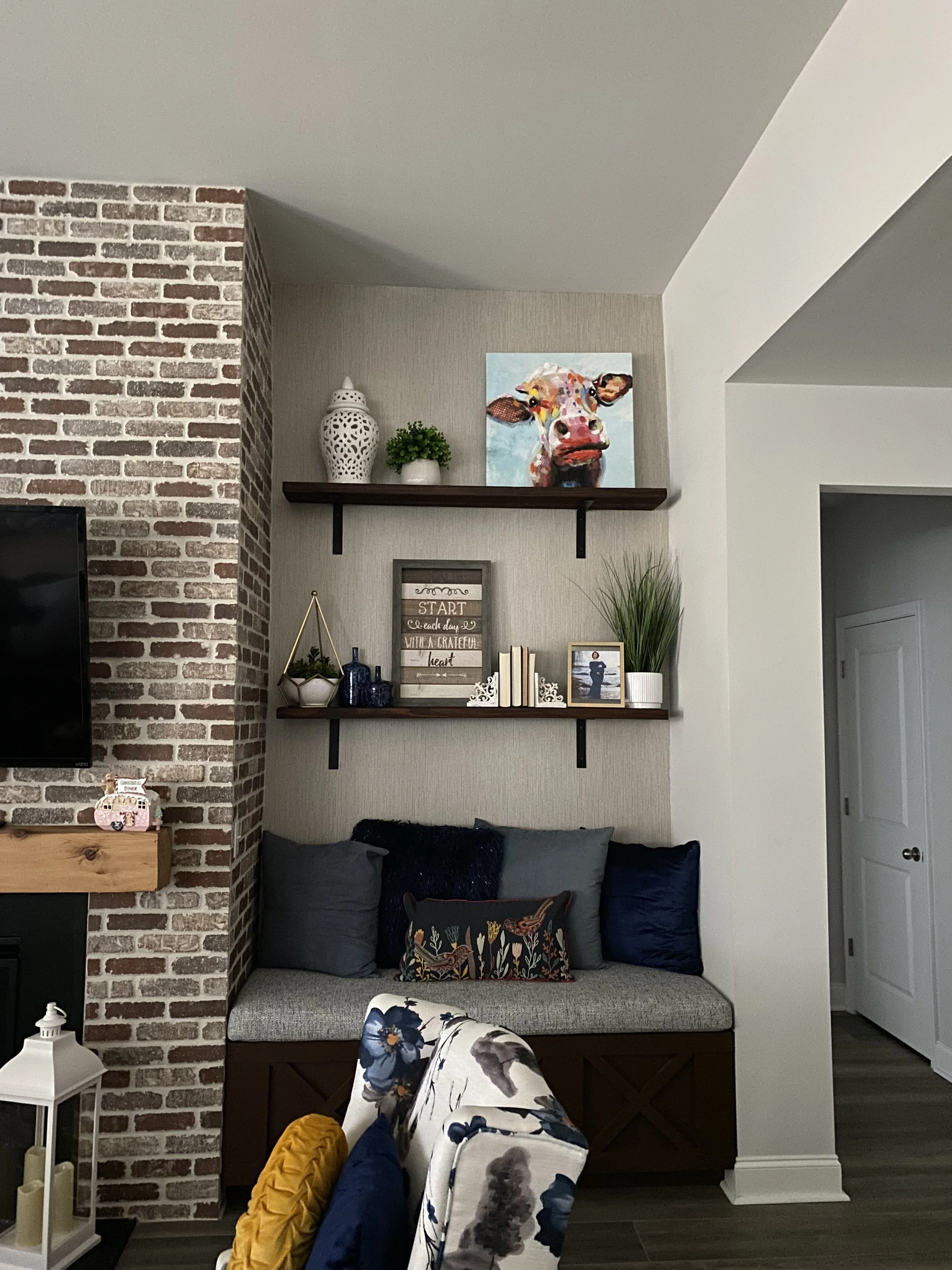 Living room corner with beige cushioned bench seat, decorative pillows, wall shelves with art and plants, brick accent wall, and a floral-patterned chair.