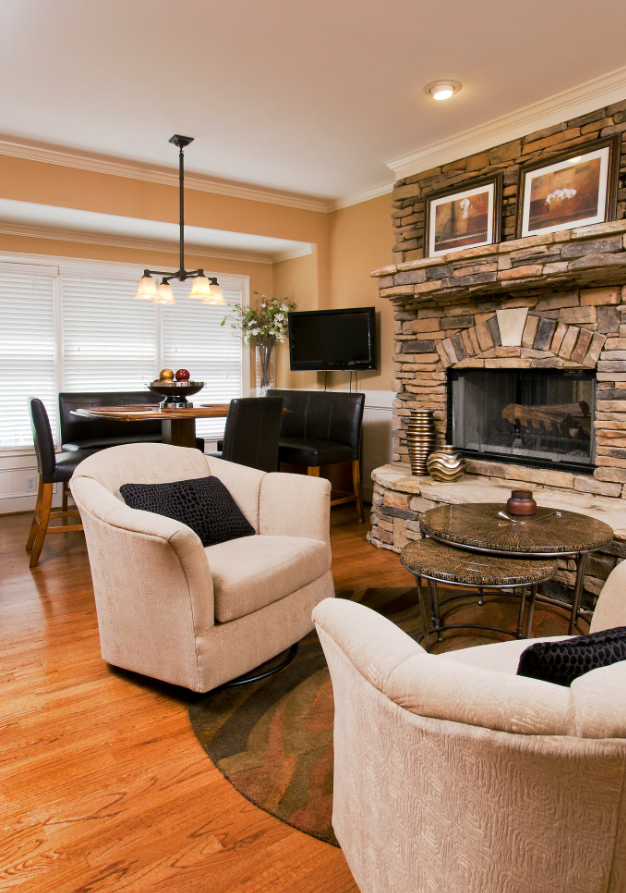 Keeping room with beige armchairs, a stone fireplace, wooden flooring, a small round table, a dining table with chairs, a wall-mounted TV, and framed artwork on the mantel
