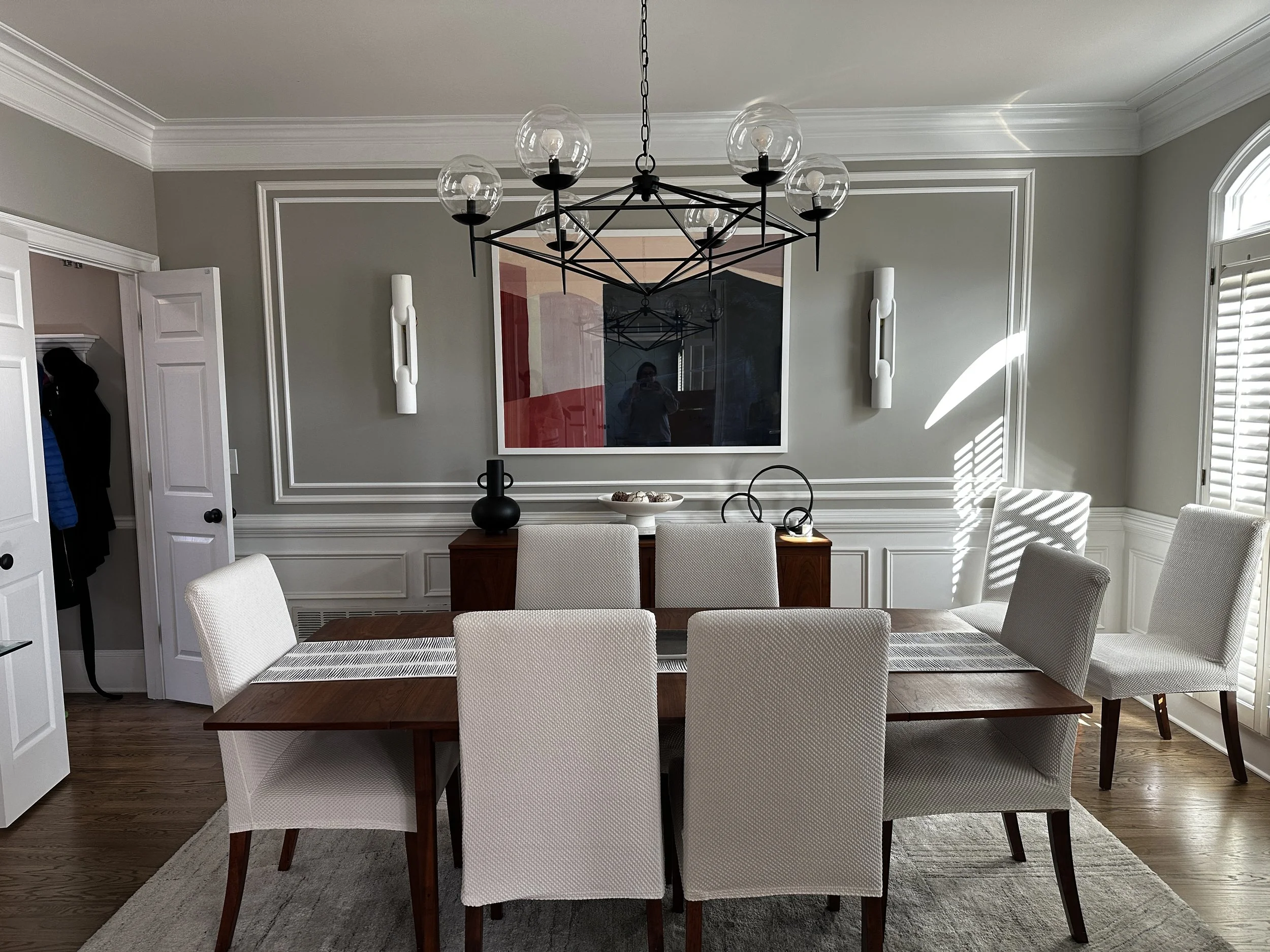 A dining room with a wooden table and six white upholstered chairs around it. The walls are painted light gray with white crown molding and wainscoting. There is a black and glass chandelier hanging above the table, and wall-mounted white light fixtu