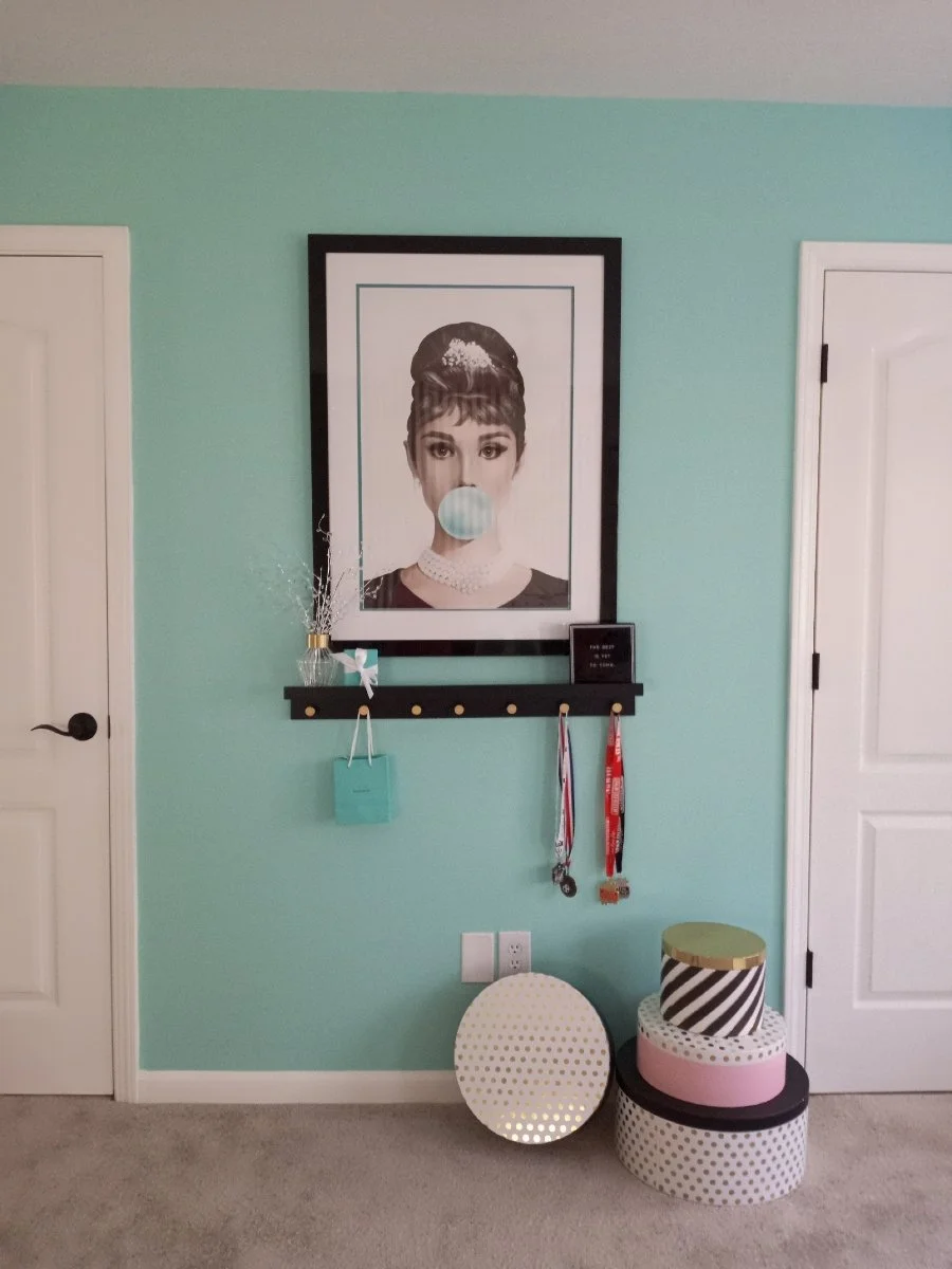 Decorative picture frame with black and white portrait of a woman with bubble gum, mint-green wall, hanging medals, a small gift bag, and stacked decorative boxes on the carpeted floor.