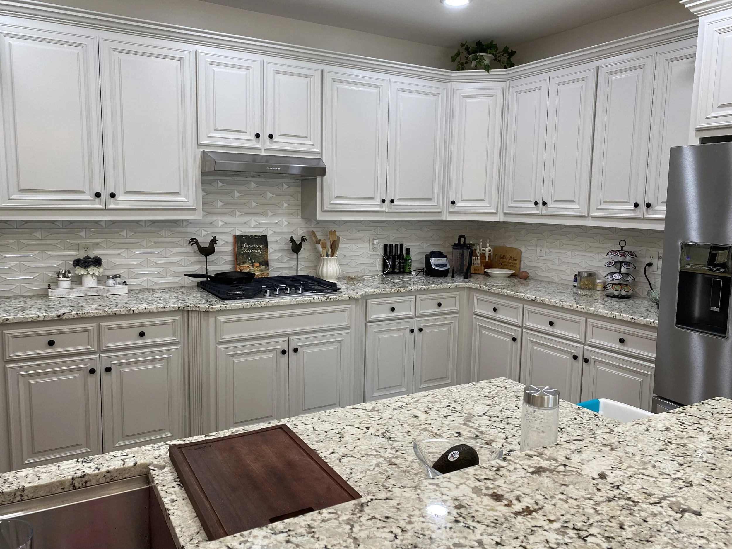 White kitchen with granite countertops, white cabinetry, and a tiled backsplash. Various kitchen utensils and appliances are visible on the counters, including a blender and a coffee maker. A wooden cutting board and decorative rooster figurines are 