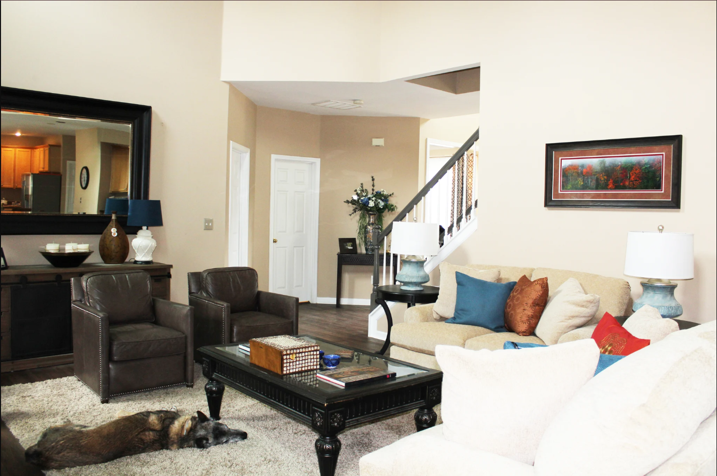 Living room with beige walls, a beige sofa with pillows, a glass coffee table, leather armchairs, table lamps, and wall artwork. A dog is lying on a rug, and a staircase with a floral arrangement is visible in the background.