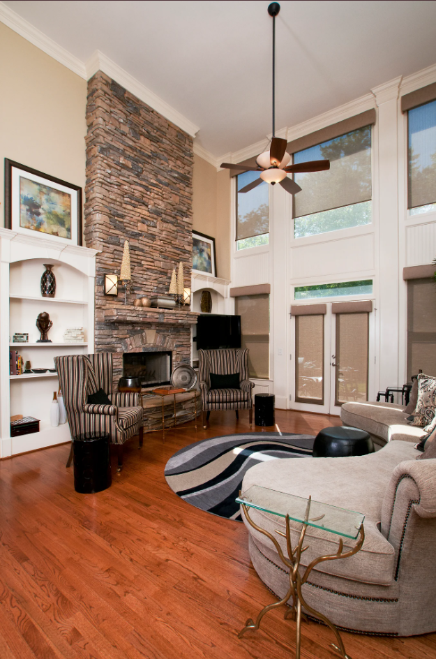 Living room with a stone fireplace, large windows with beige blinds, striped armchairs, a beige sectional sofa, a black ottoman, a patterned area rug, and decorative shelves with artwork.