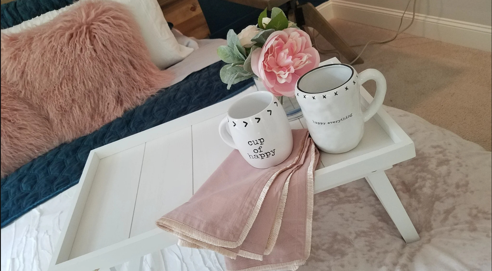A white tray table on a bed with a pink cloth napkin, two white mugs with black lettering and designs, a pink flower in a white vase, and a pink fluffy pillow on the bed in the background.