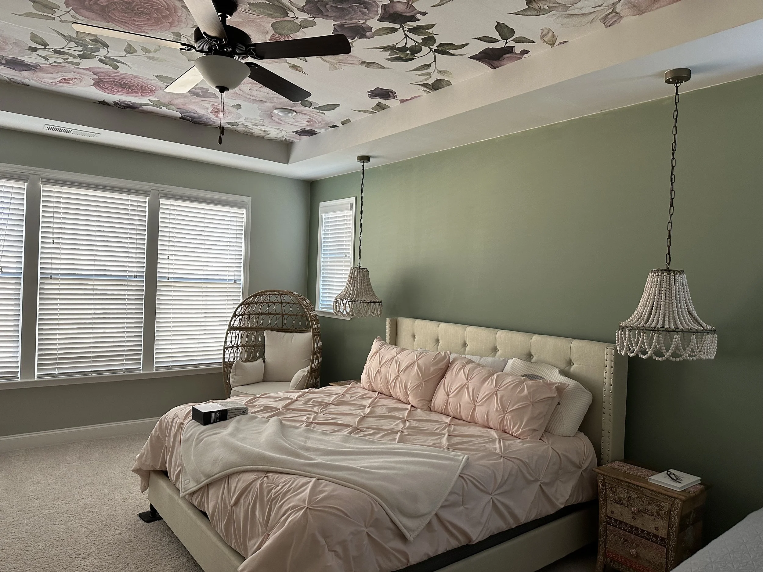 A bedroom with a bed, pink quilt, and multiple throw pillows. There are two hanging lamps with beaded shades and a wicker chair. Large windows with white blinds are on the wall opposite the bed. The ceiling has a floral pattern with roses and green l