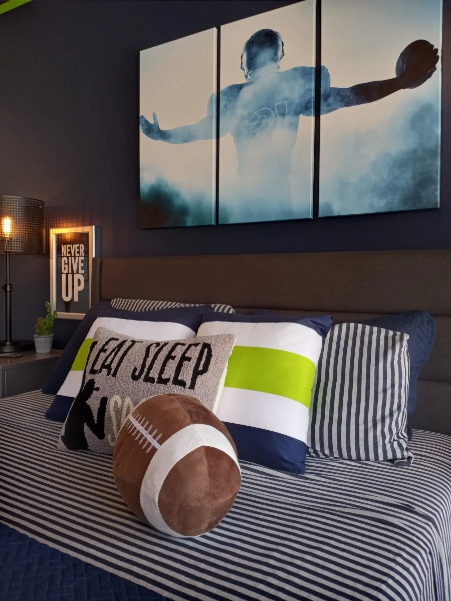 Bedroom with striped bedspread, pillows including one with "Eat Sleep" text, football-shaped pillow, side table, lamp, and sports-themed wall art featuring a player with outstretched arms holding a football.