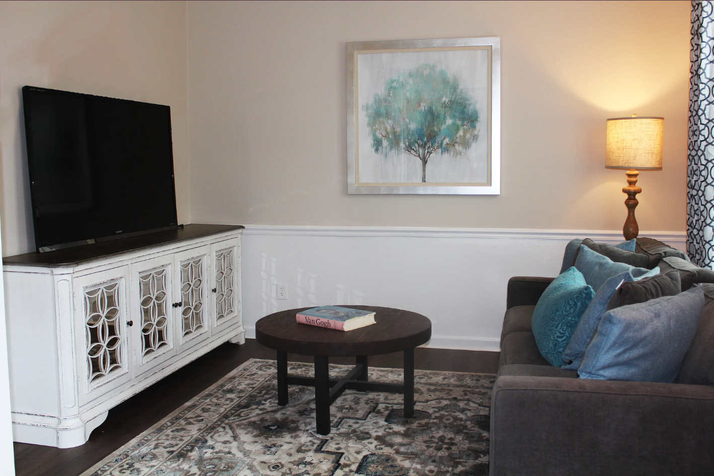 Living room with a flat-screen TV on a white cabinet, a dark wooden coffee table with a book, a gray sofa with blue pillows, a framed picture of a tree, a lamp, and a patterned curtain.