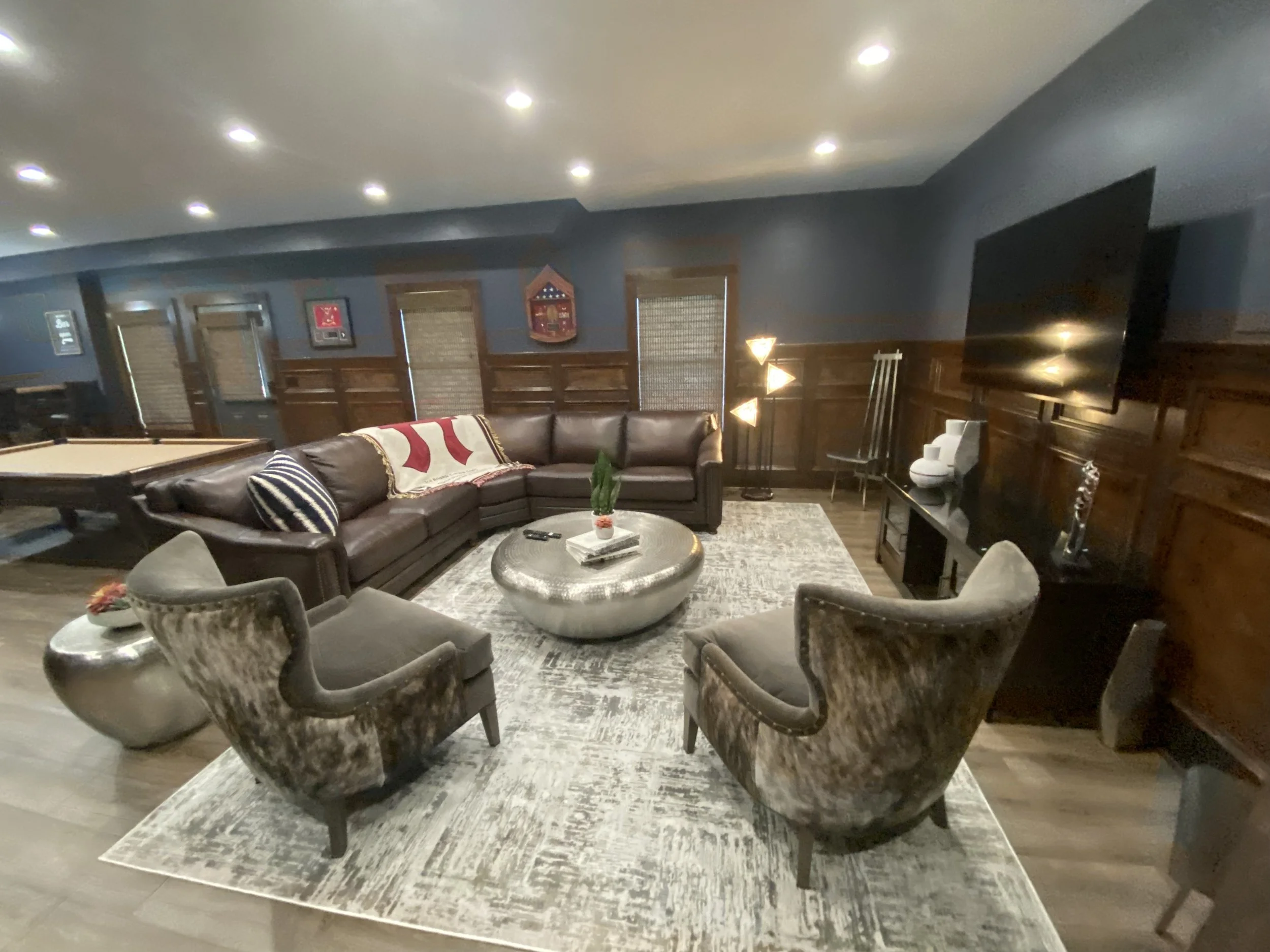 Basement living room with dark wood paneling, a leather sectional sofa with pillows, a coffee table, a TV, and decorative lighting.