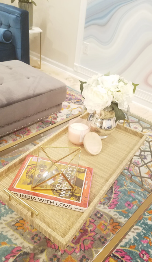 Decorative living room coffee table with flowers, candles, books, and a glass geometric sculpture, with a colorful rug and part of a gray sofa in the background.