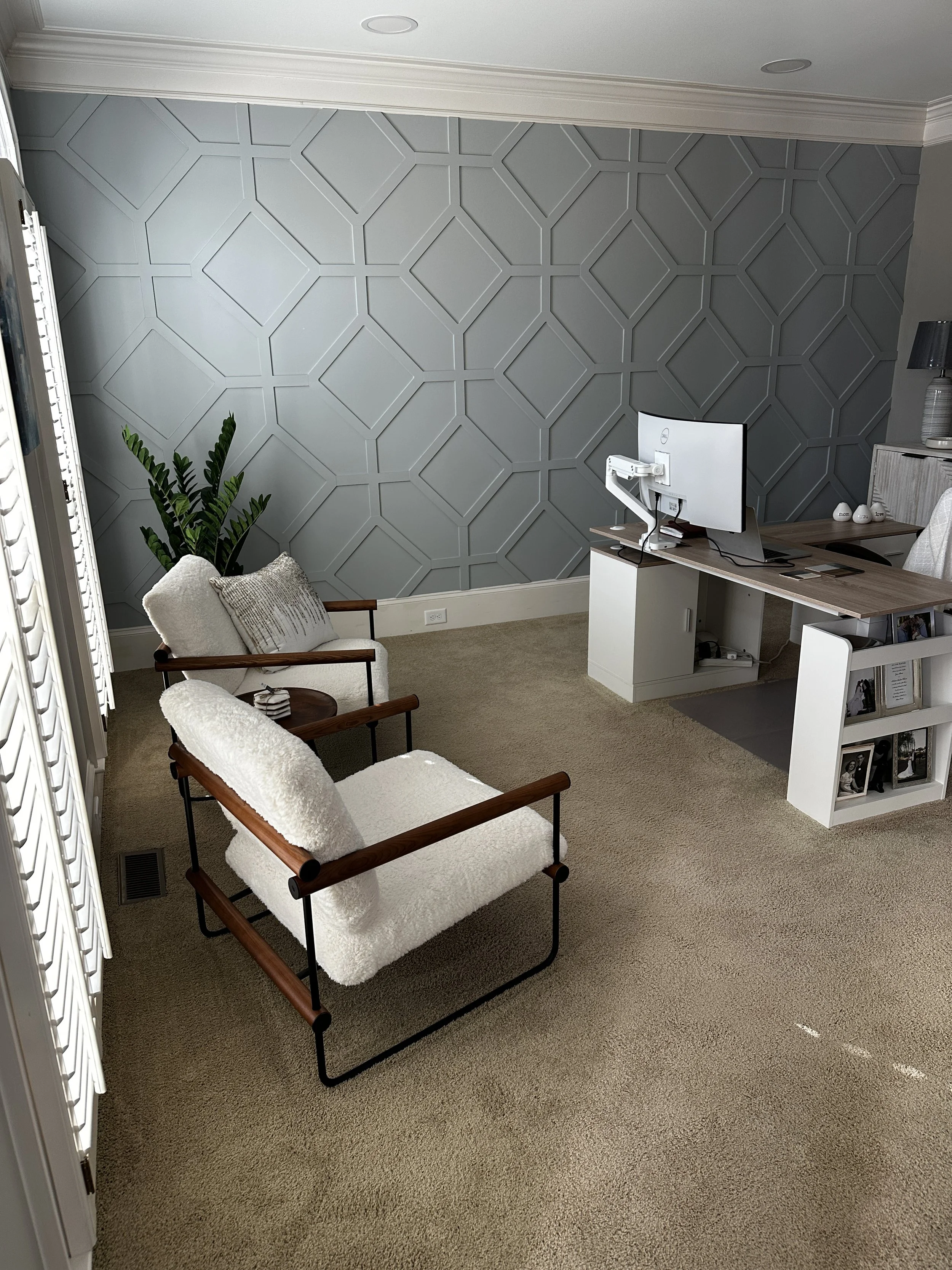 A home office with a patterned gray accent wall, two mid-century modern style armchairs with white cushions, a green indoor plant, a white desk with a monitor, keyboard, and mouse, and a white shelving unit with framed photos and decor. A lamp and de