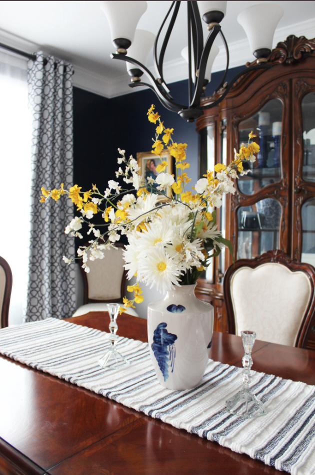 A dining room table with a striped table runner, a vase with white and yellow flowers, and two small glass candle holders. A chandelier hangs above, and a china cabinet with glass doors is in the background.