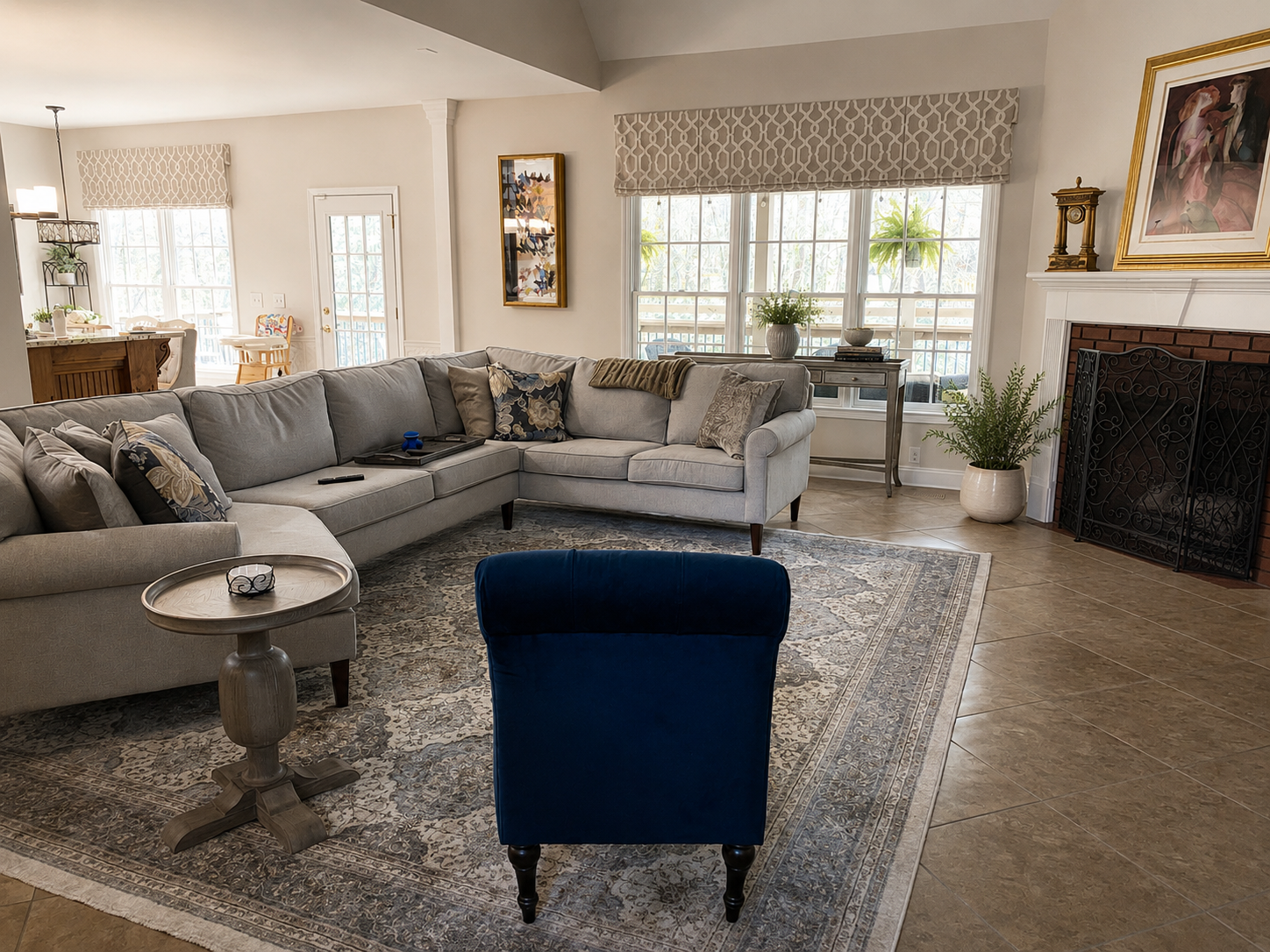 A living room with a large light gray sectional sofa decorated with multiple pillows, a dark blue armchair, a round side table, and a patterned area rug. The room has large windows with valances, a fireplace with a brick facade, and houseplants. Ther