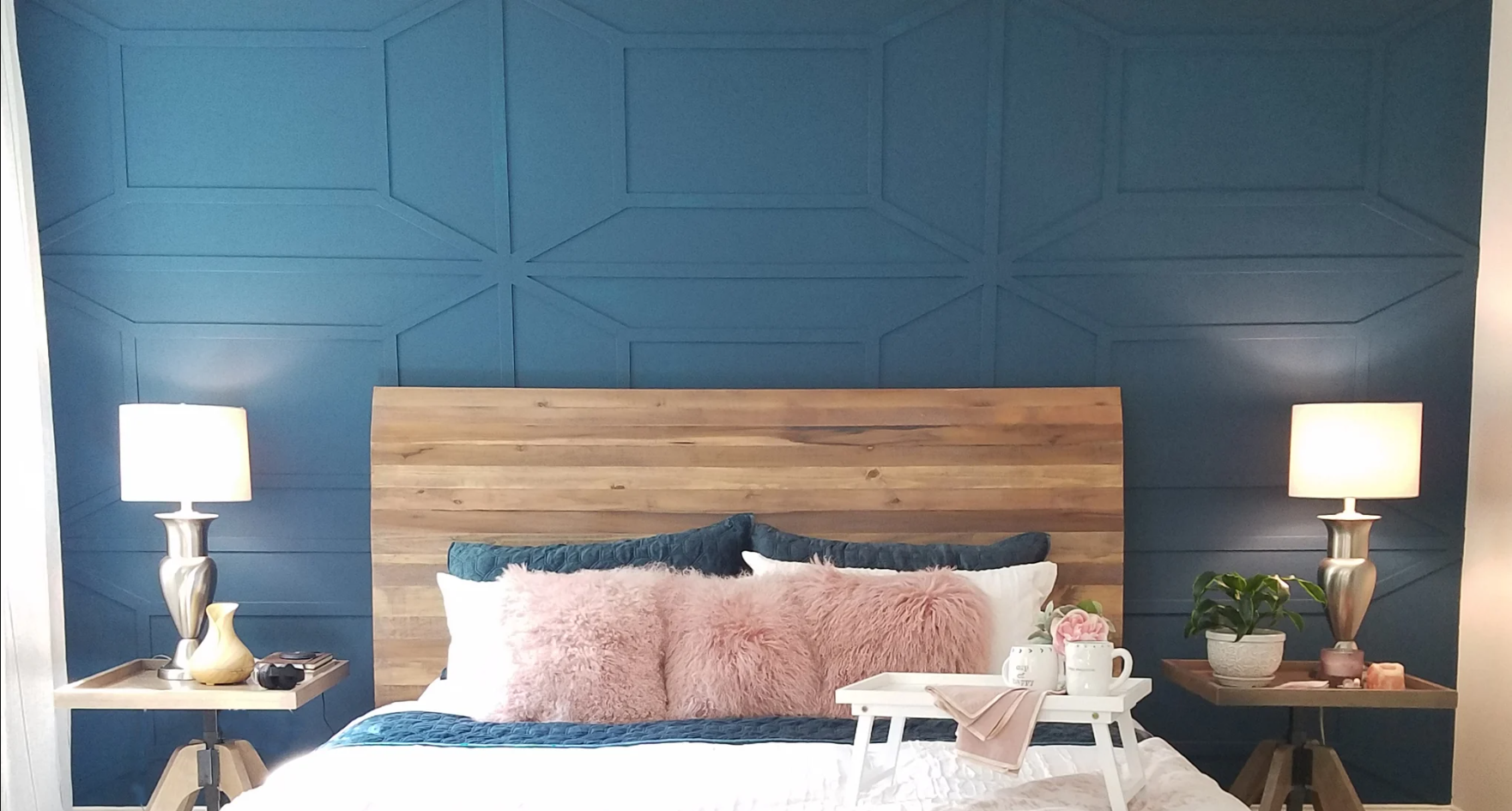 A cozy bedroom features a wooden headboard against a deep blue wall with geometric paneling. Two bedside tables hold lamps, a plant, and decorative items. The bed is decorated with pink and blue pillows and a white blanket with a tray holding mugs an