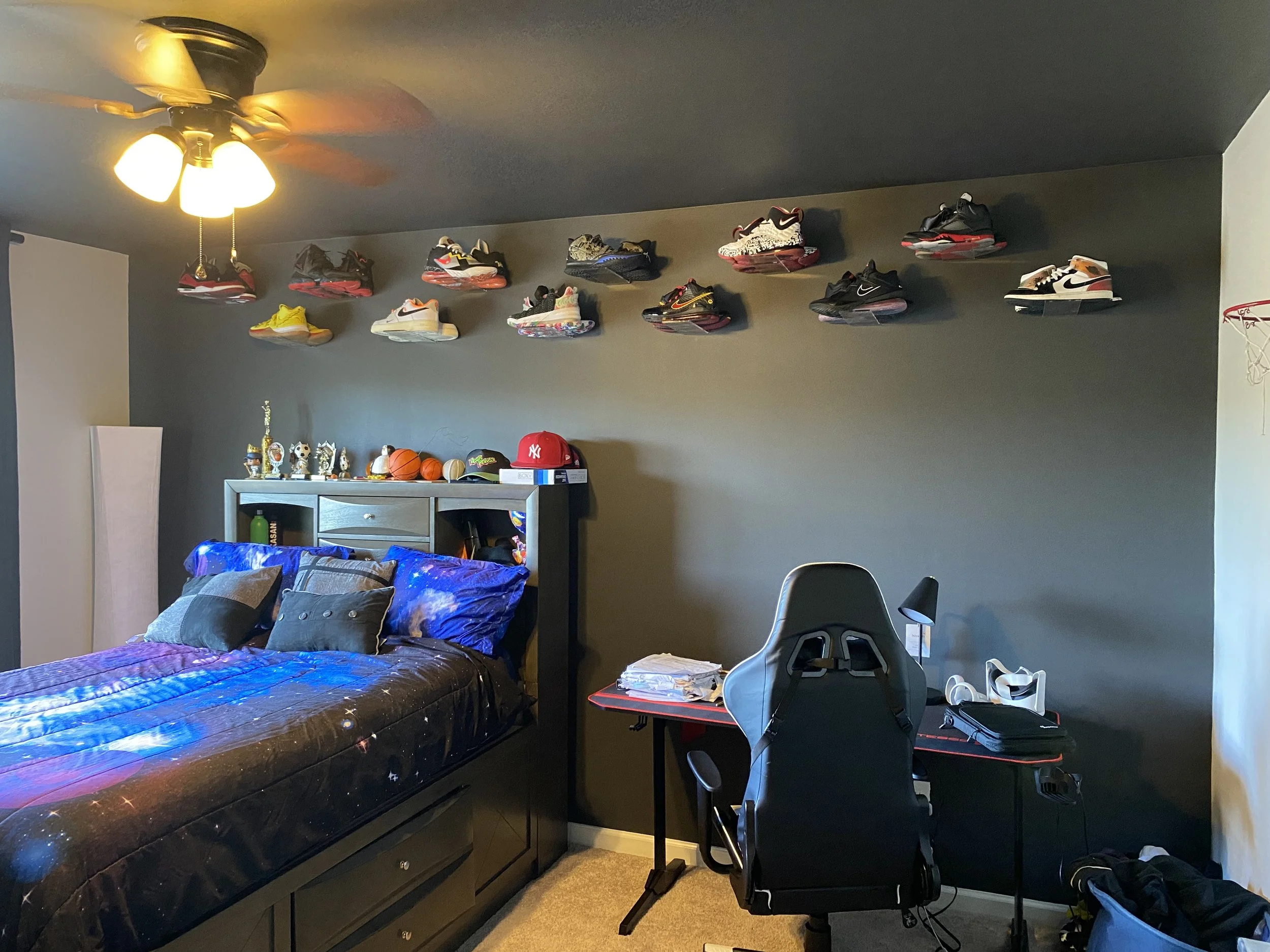 A bedroom with a bed, a ceiling fan with lights, a shelf with trophies and basketballs, and a desk with a computer chair, notebooks, and other items. The wall behind the bed is painted dark gray and displays athletic shoe collection.