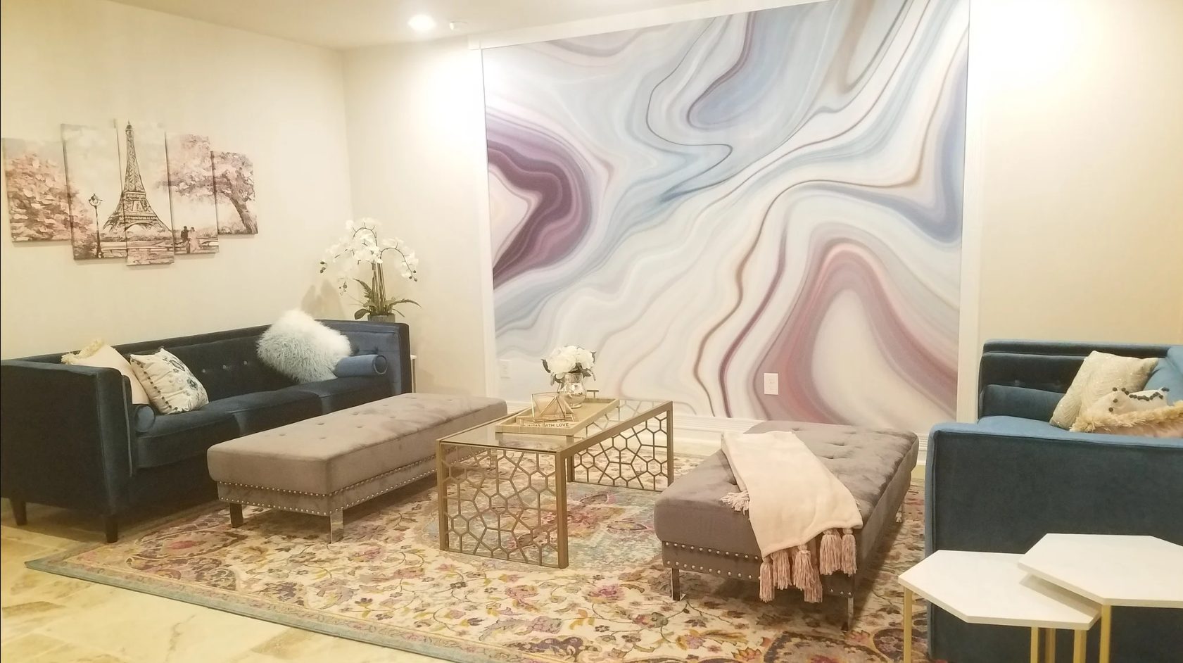 Living room with two dark couches, upholstered ottomans, a glass coffee table, a floral area rug, wall art, a decorative wall with swirling abstract design, and various decorative pillows and plants.
