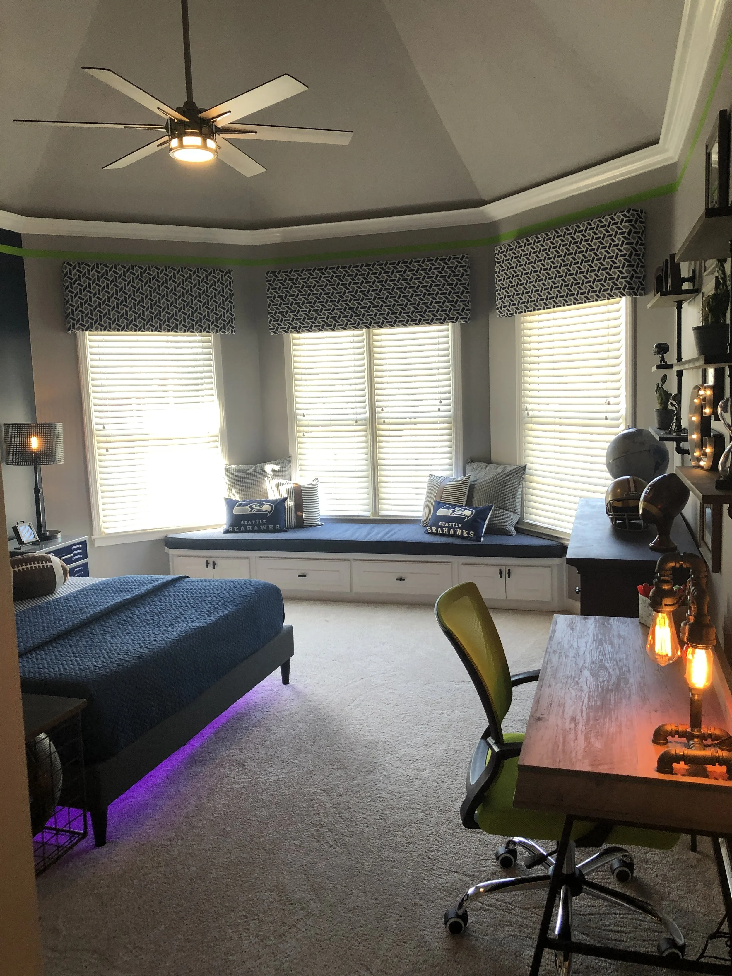 A young boy's bedroom with a window seat under three large windows with patterned window valances, a ceiling fan, and a workspace with a wooden desk and green ergonomic chair, decorated with lamps, pillows, and various decorative items.