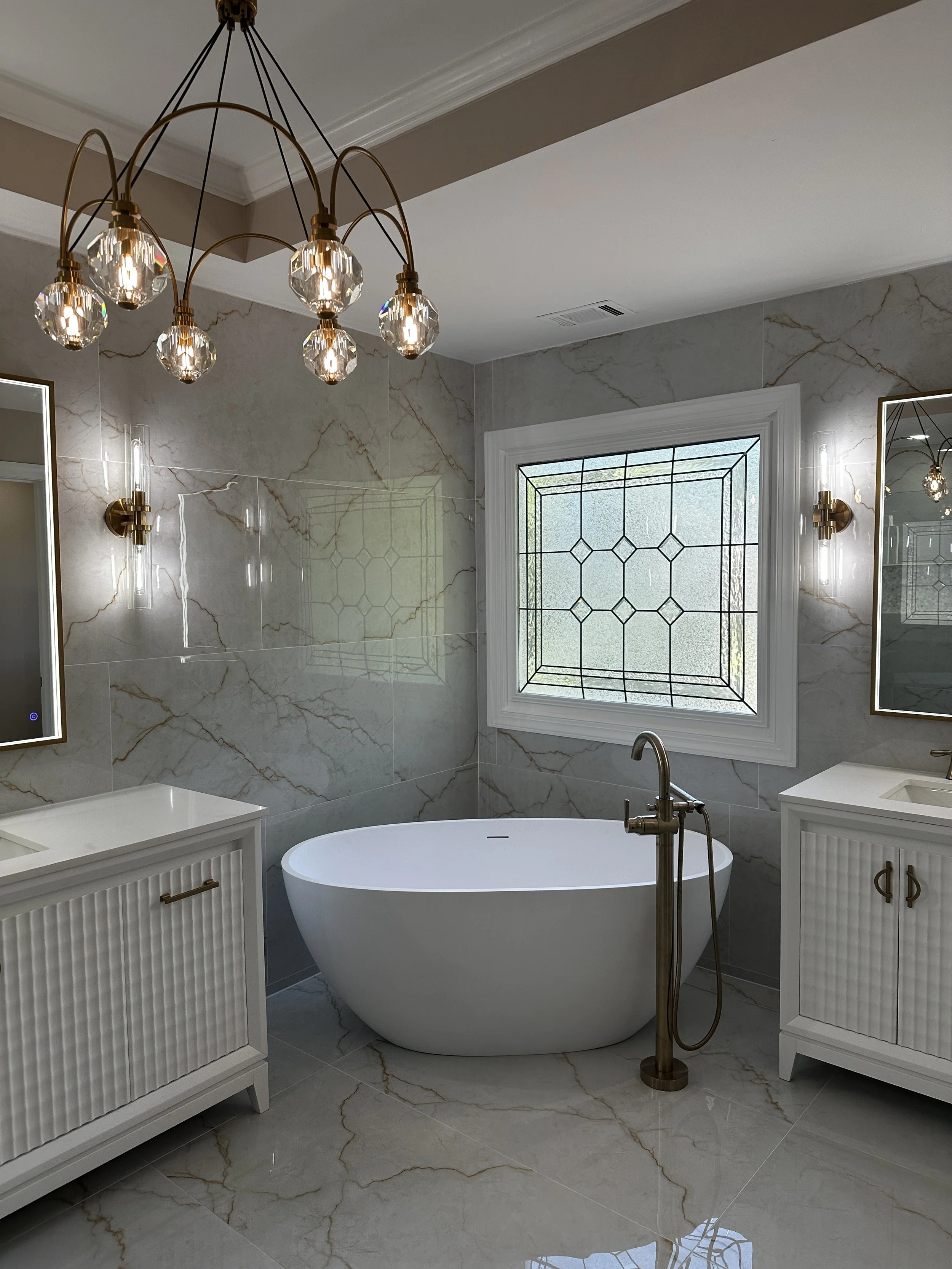 Modern bathroom with a white free-standing bathtub, marble tile walls and floor, a window with decorative metal grille, and two matching framed mirrors. There is a brass chandelier with crystal accents and brass wall sconces.